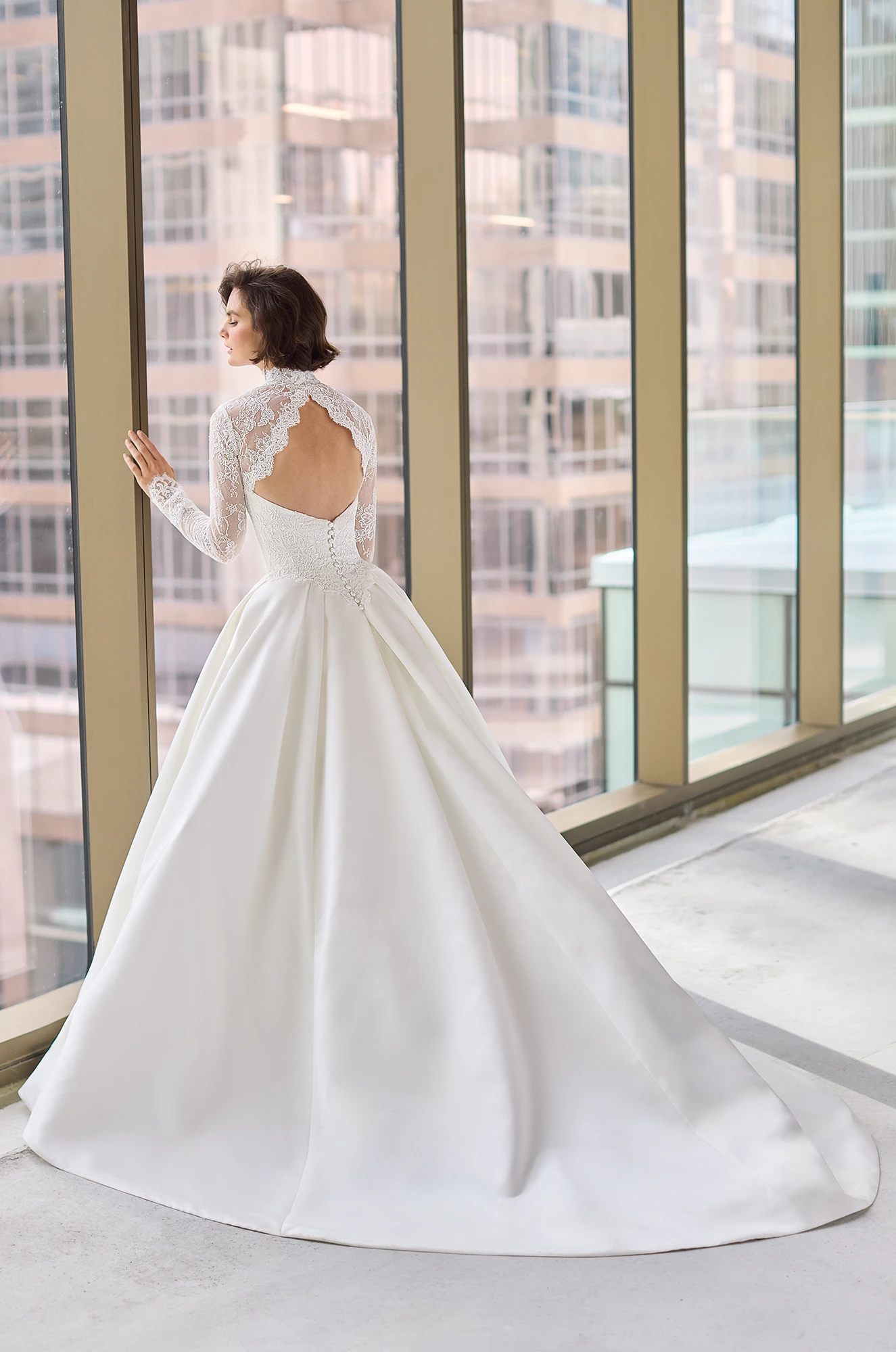 Back view of a wedding dress with a Lace bodice and long sleeve Lace bolero with a keyhole back, and a pleated Satin ball gown skirt, in front of large windows overlooking a city.