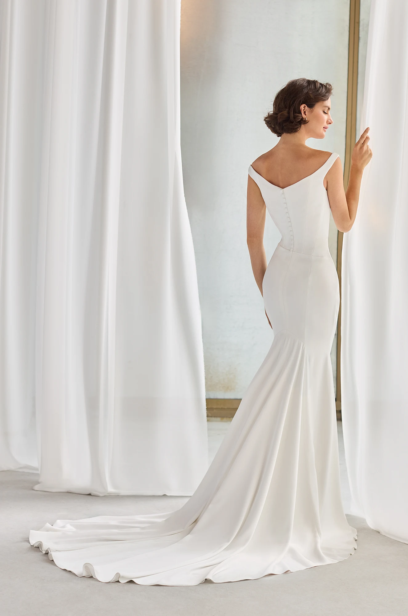 Back view of an off the shoulder Charmeuse wedding dress with an asymmetric waistline and a fit and flare skirt, in a studio with white flowing curtains.