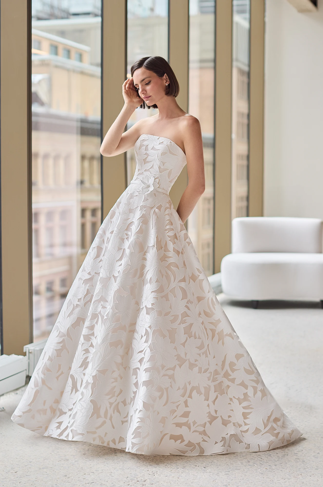 Front view of a strapless Lace wedding dress with a straight neckline and an A-line skirt, in front of large windows overlooking a city.