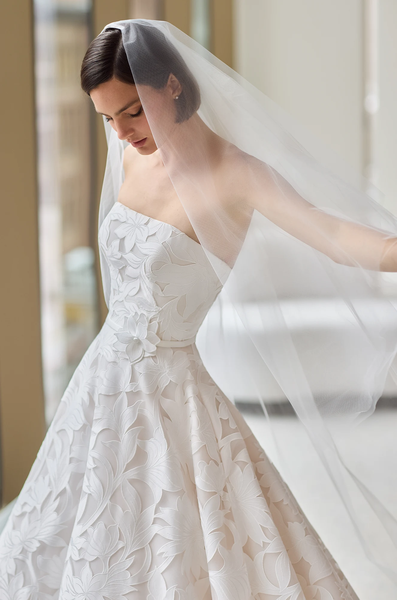 Close up view of a model wearing a strapless Lace wedding dress with a straight neckline and a veil, in a studio.