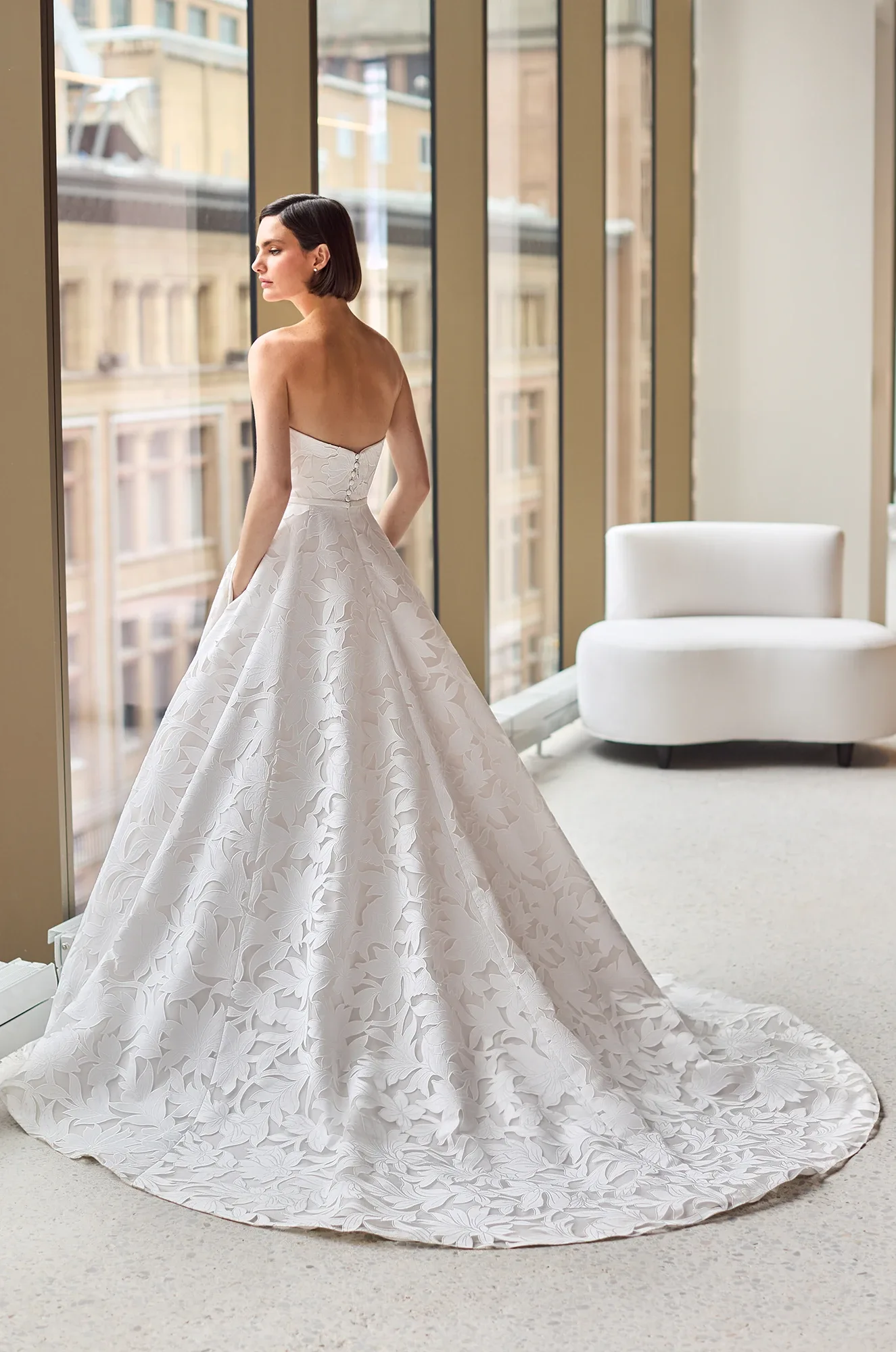 Back view of a strapless Lace wedding dress with buttons on the back of the bodice and an A-line skirt with pockets, in front of large windows overlooking a city.