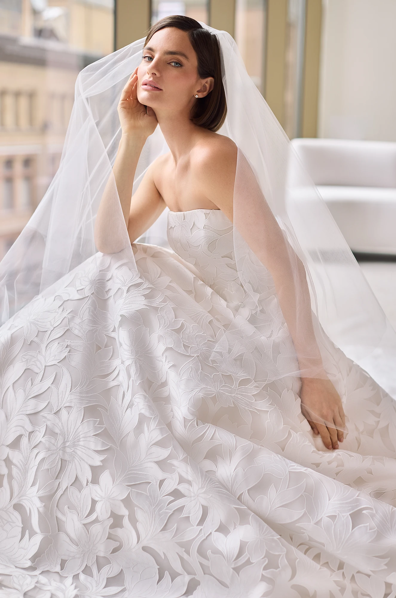 Close up view of a model wearing a strapless Lace wedding dress and a veil, sitting on the floor of a studio with large windows.