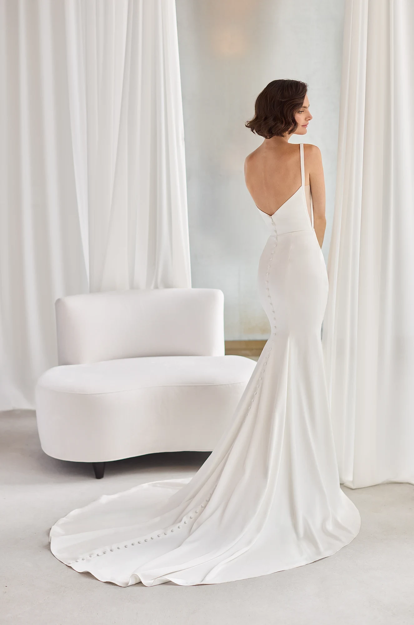 Back view of a sleeveless Charmeuse wedding dress with a fit and flare skirt that has buttons along the back of the dress down to the end of the train, in a studio with flowing white curtains and a lounge chair.