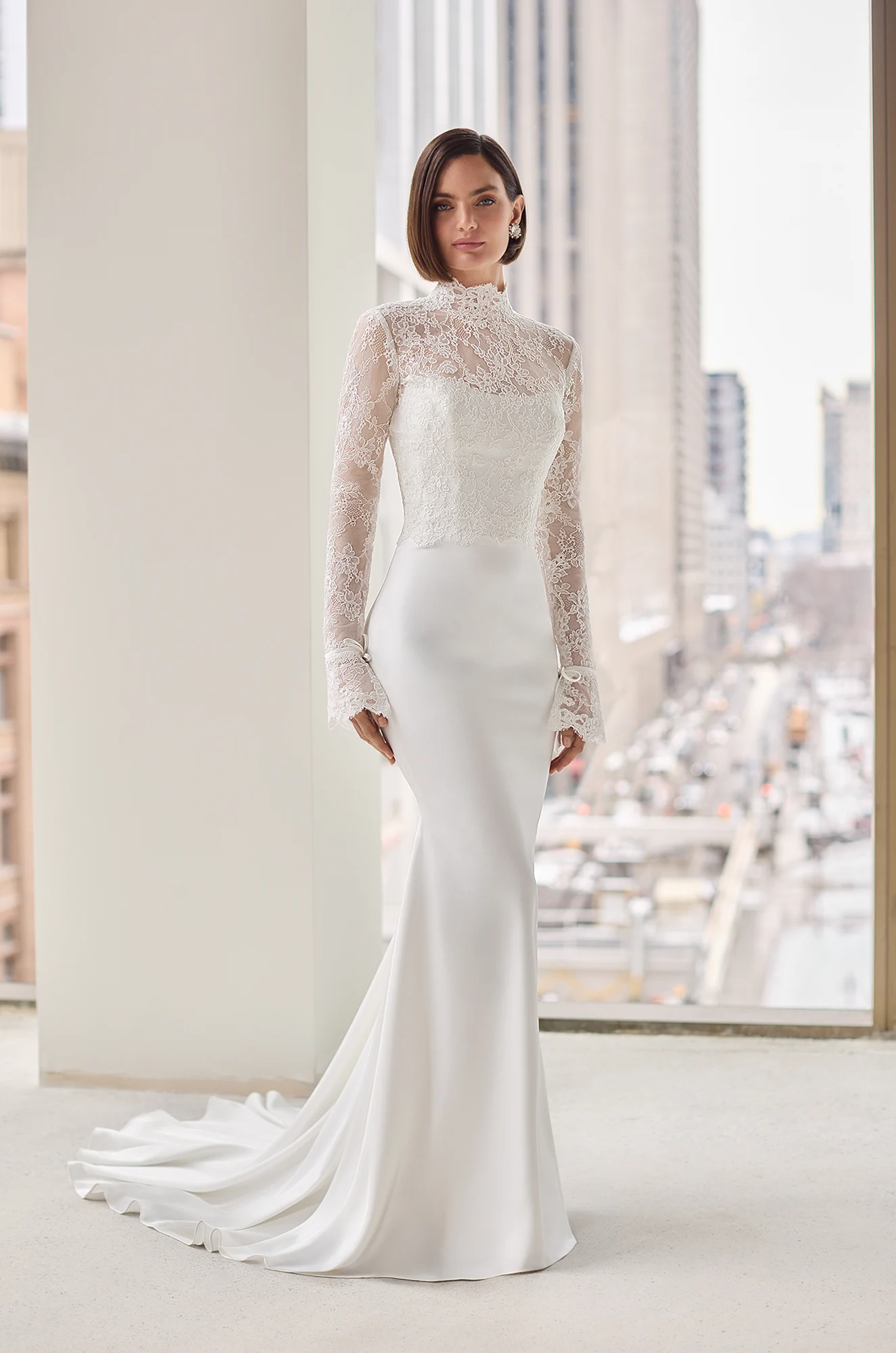 Front view of a strapless Charmeuse wedding dress with a scoop neckline and a fit and flare skirt, in front of large windows overlooking a city.