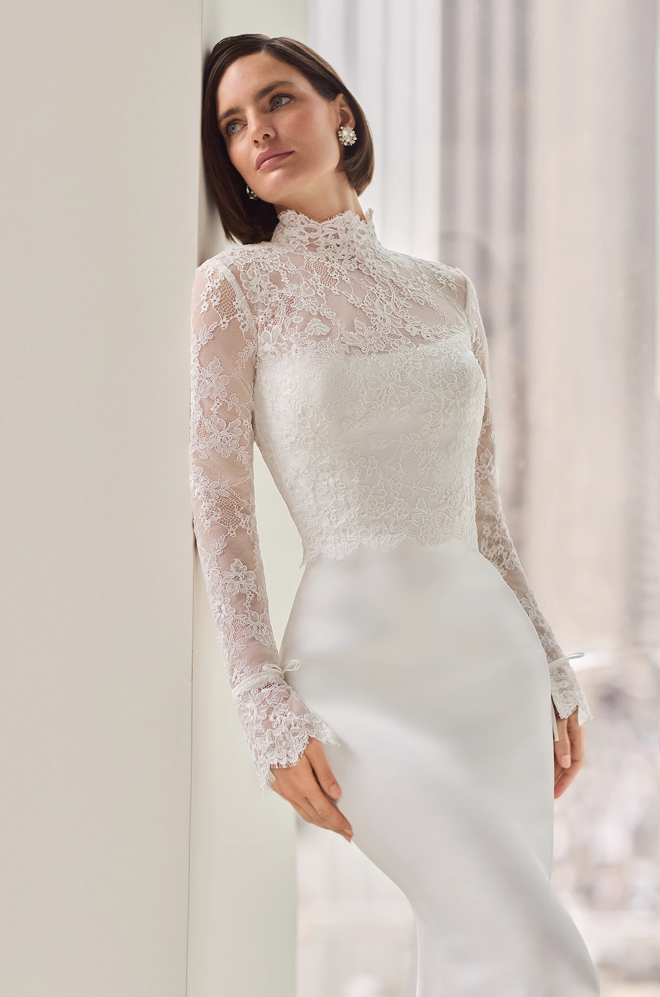 Close up view of a strapless Charmeuse wedding dress with a fitted skirt and a long sleeve sheer Lace topper over the bodice of the dress, in front of a window overlooking a city.