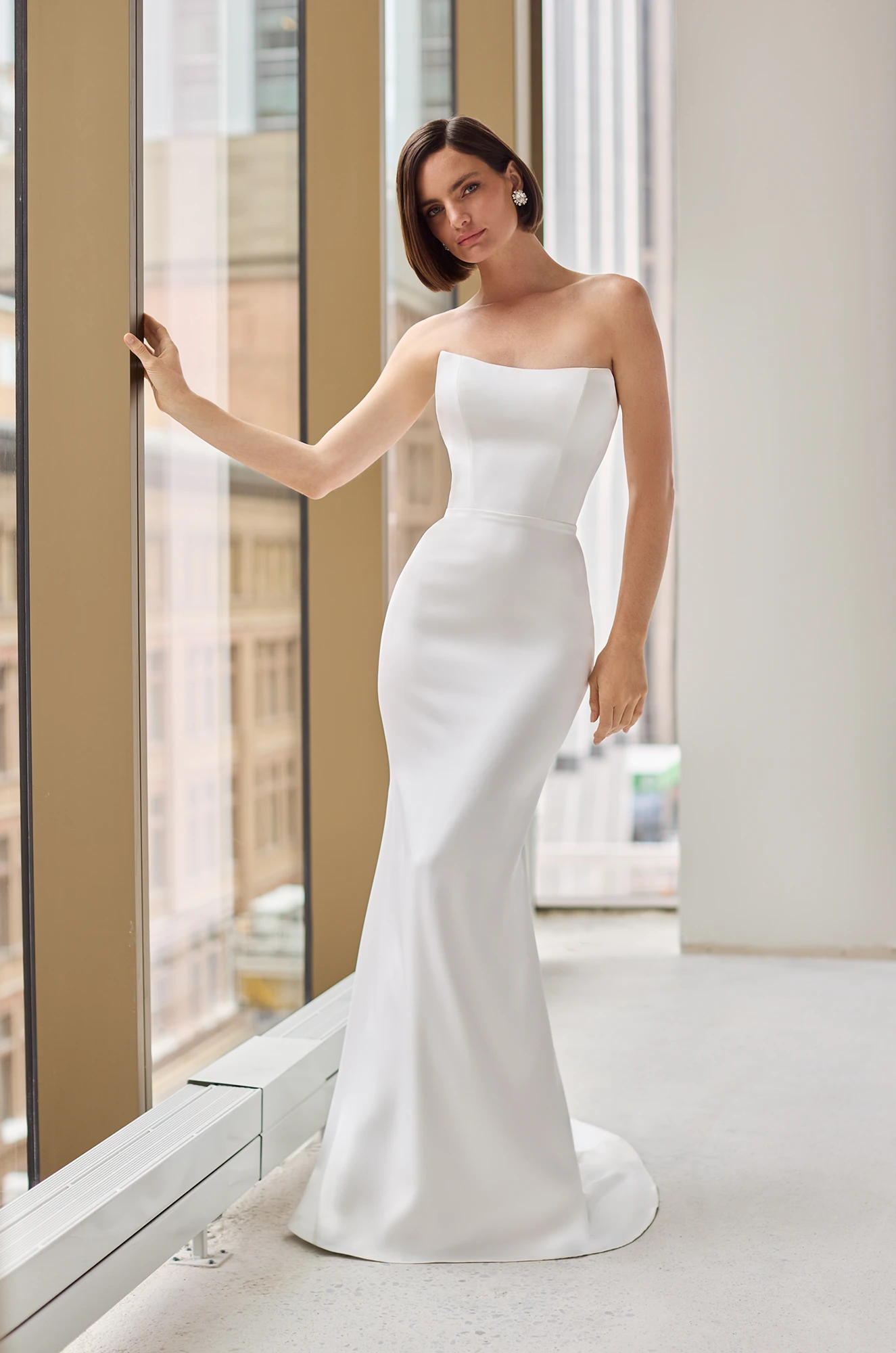 Front view of a strapless Charmeuse wedding dress with a fitted skirt and a long sleeve sheer Lace topper over the bodice of the dress, in front of a window overlooking a city.