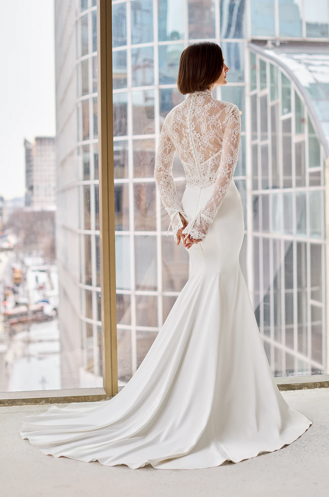Back view of a strapless Charmeuse wedding dress with a fit and flare skirt and a long sleeve sheer Lace topper over the bodice of the dress, in front of a window overlooking a city.