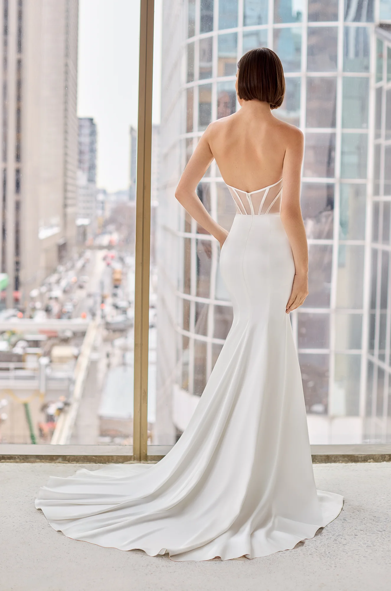 Back view of a strapless Charmeuse wedding dress with a sheer corset back and a fit and flare skirt, in front of a window overlooking a city.