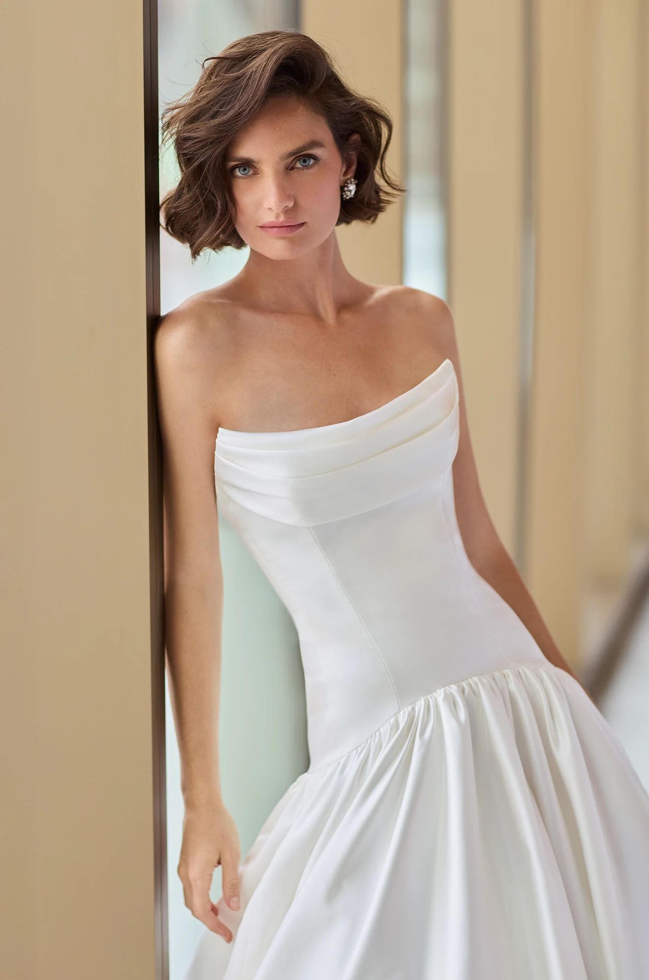 Close up view of a strapless Satin wedding dress with a draped scoop neckline and drop waisted bodice, with a gathered skirt, in front of windows.