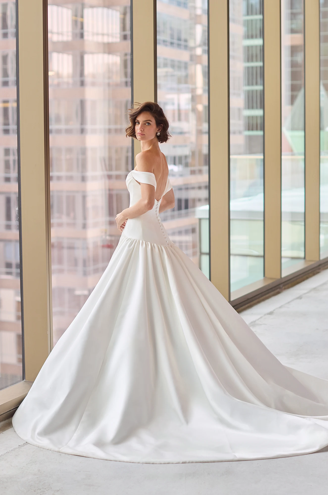Back view of a strapless Satin wedding dress with draped sleeves, an open back, and a drop waisted bodice, with a gathered ball gown skirt, in front of large windows overlooking a city.