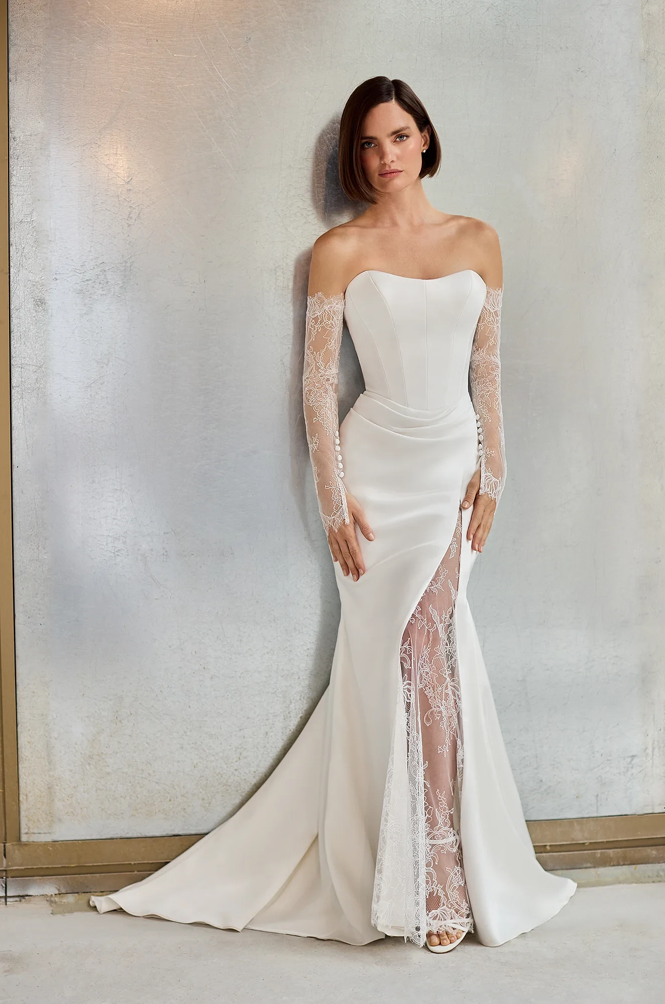 Front view of a strapless Charmeuse wedding dress with a fit and flare skirt and a Lace insert in the skirt slit, with separate long Lace sleeves, in front of a wall.