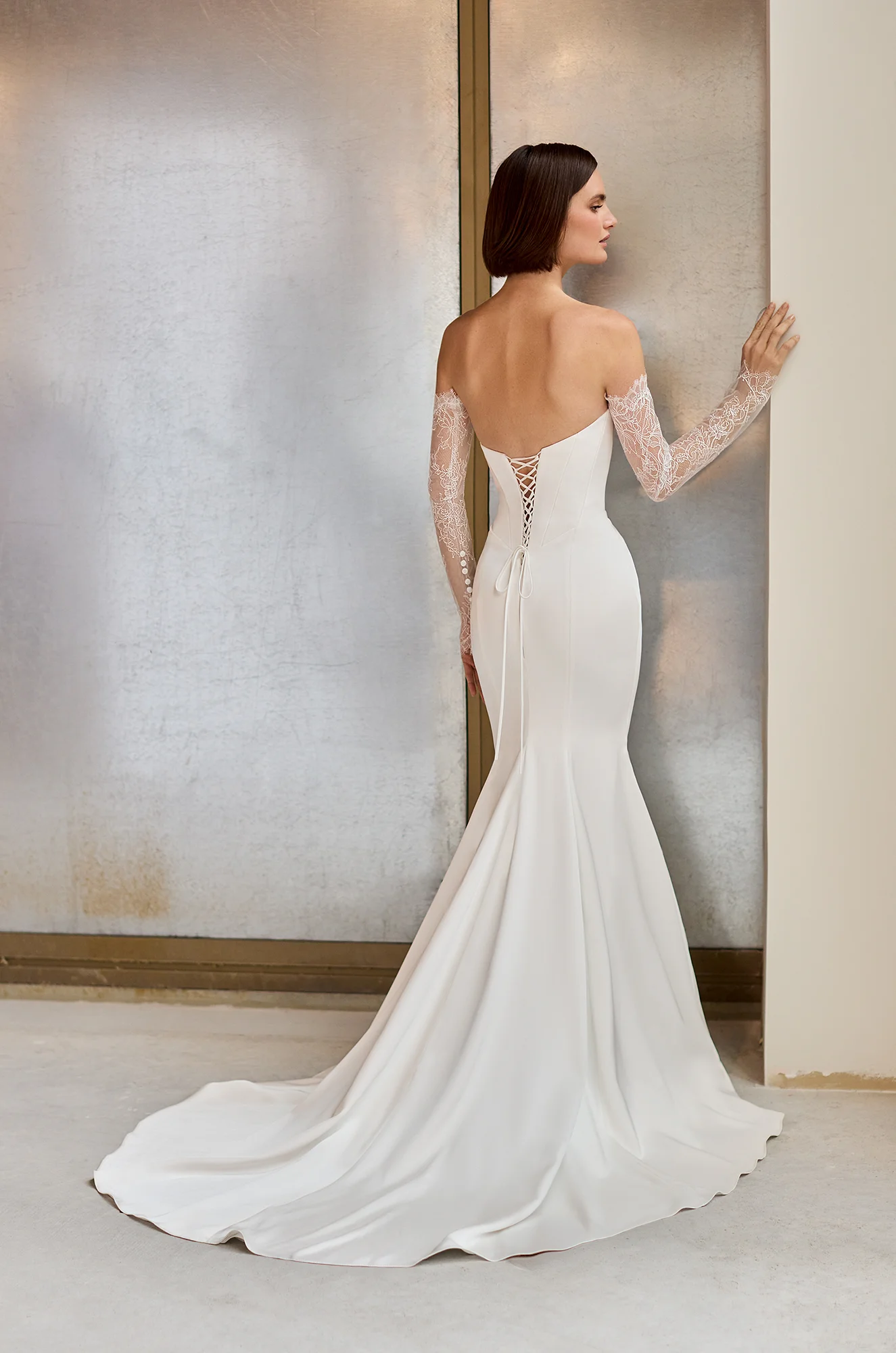Back view of a strapless Charmeuse wedding dress with a lace-up back closure, a fit and flare skirt, and separate long Lace sleeves, in front of a wall.