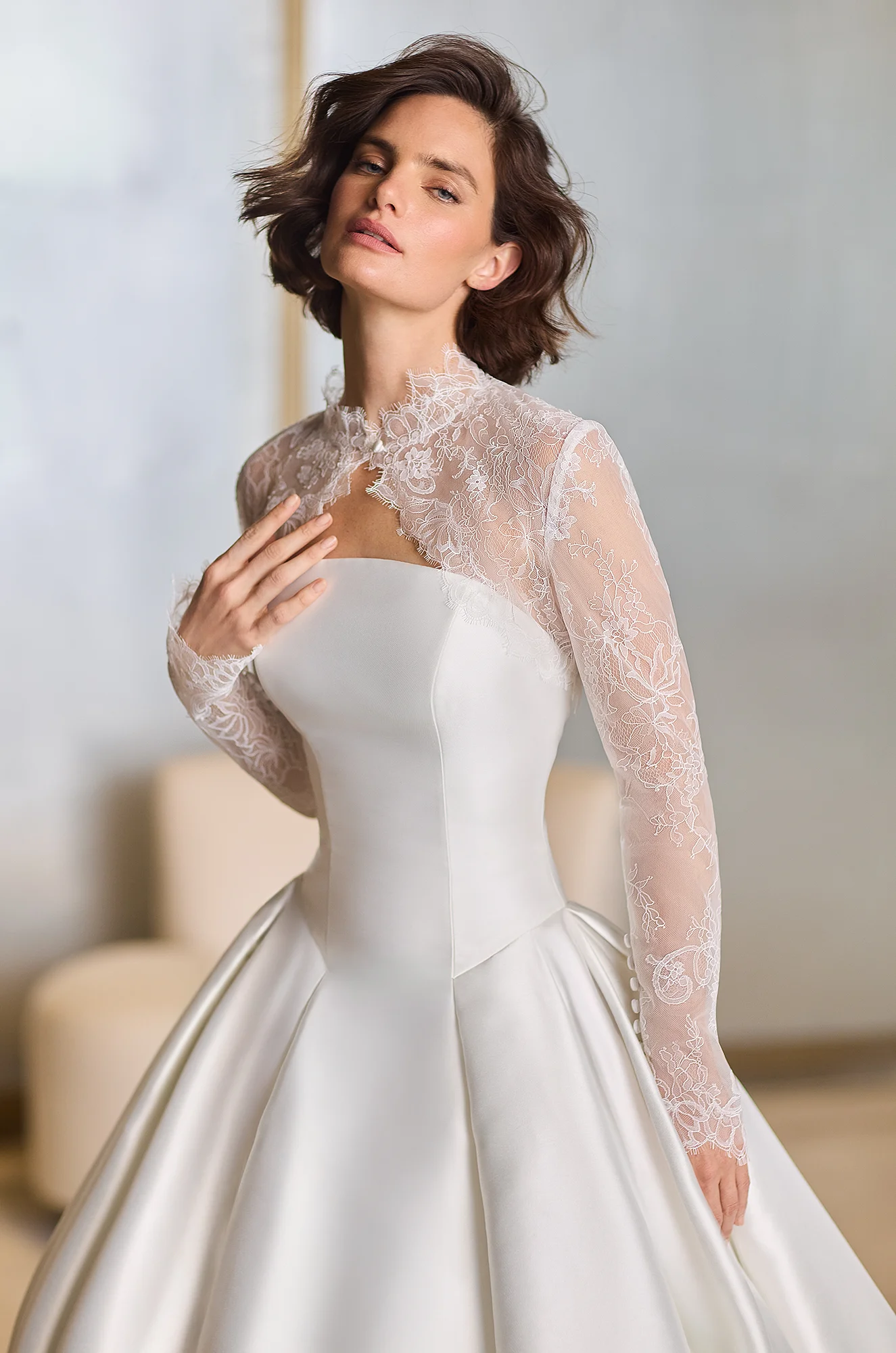 Close up view of a strapless Mikado princess cut wedding dress with an arched neckline and box pleated ball gown skirt, with a removable long sleeve sheer Lace bolero, in a studio.