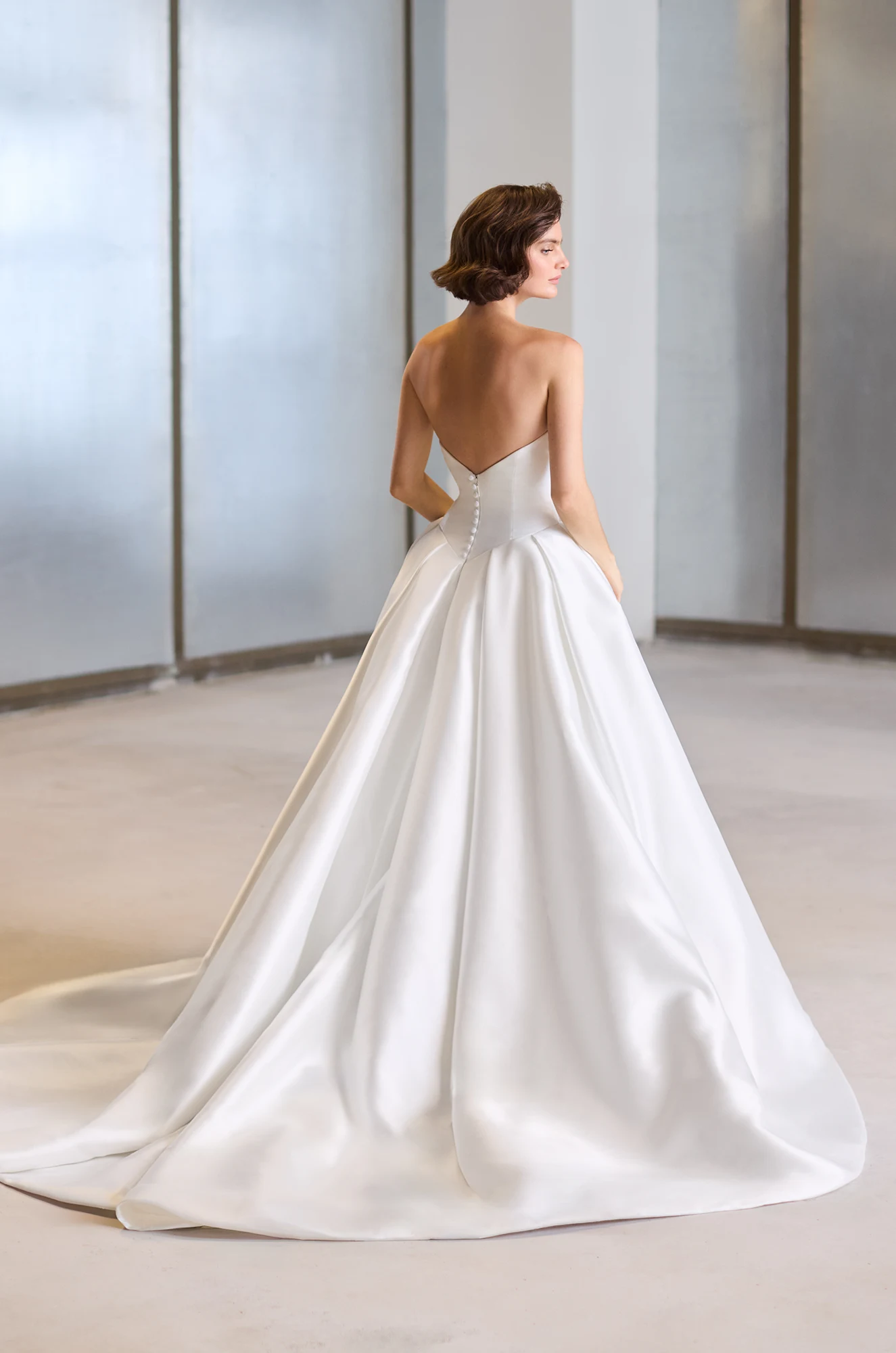 Back view of a strapless Mikado wedding dress with buttons on the back of the bodice and a box pleated ball gown skirt, in a studio.