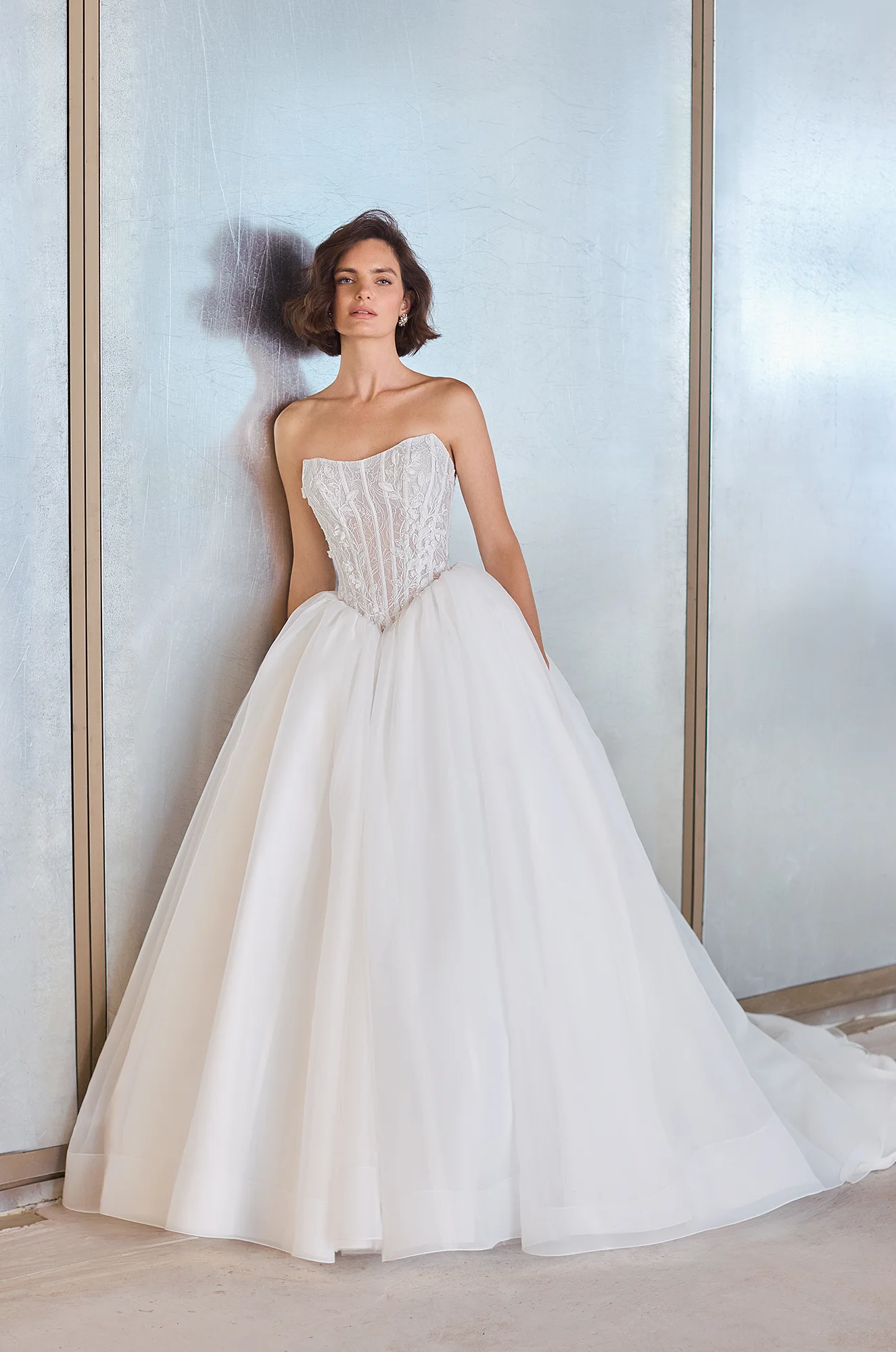 Front view of a strapless sheer Lace bodice wedding dress with a basque waistline and a gathered Organza ball gown skirt, in front of a wall.