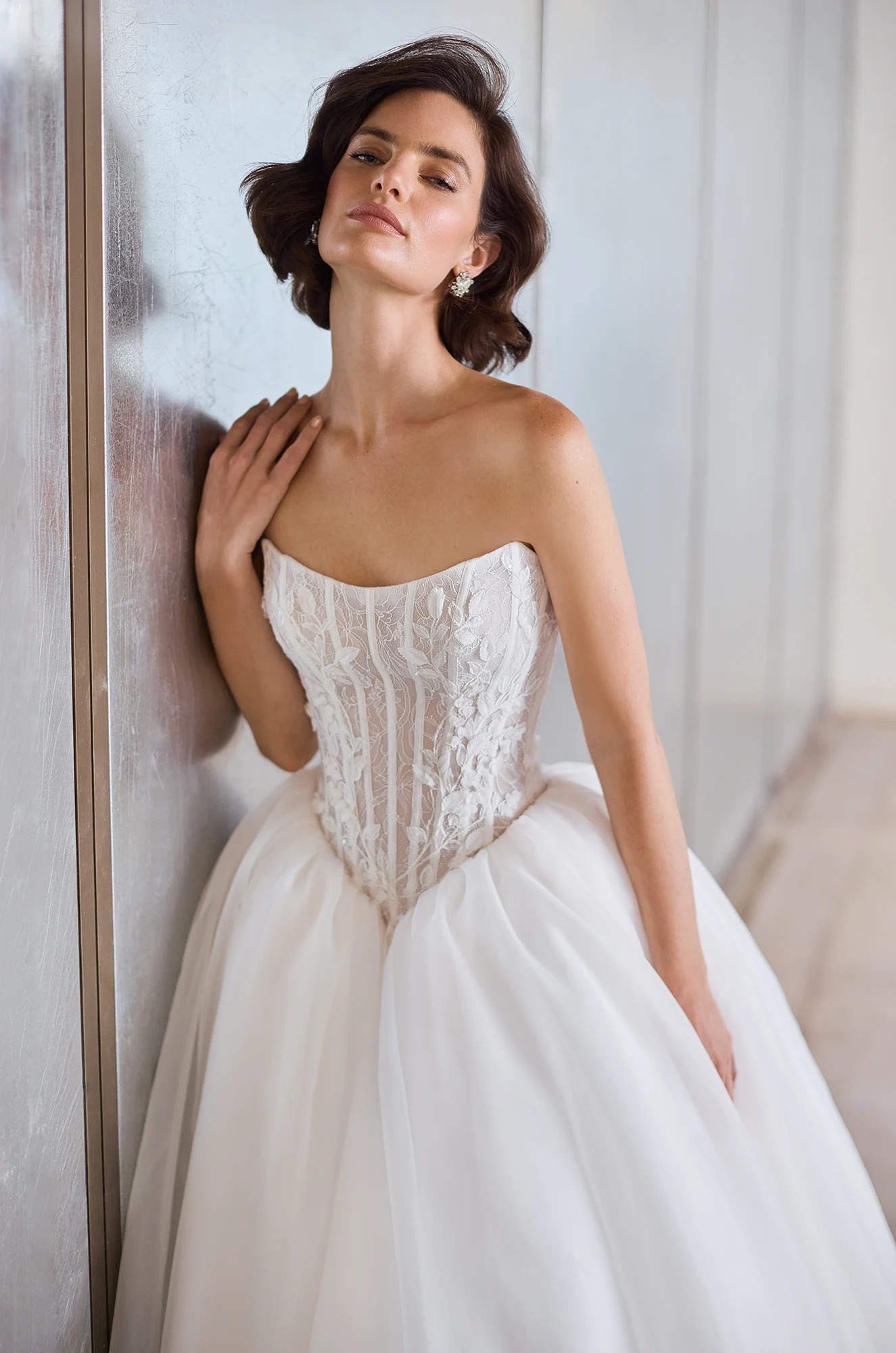 Close up view of a strapless sheer Lace bodice wedding dress with a basque waistline and a gathered Organza ball gown skirt, in front of a wall.