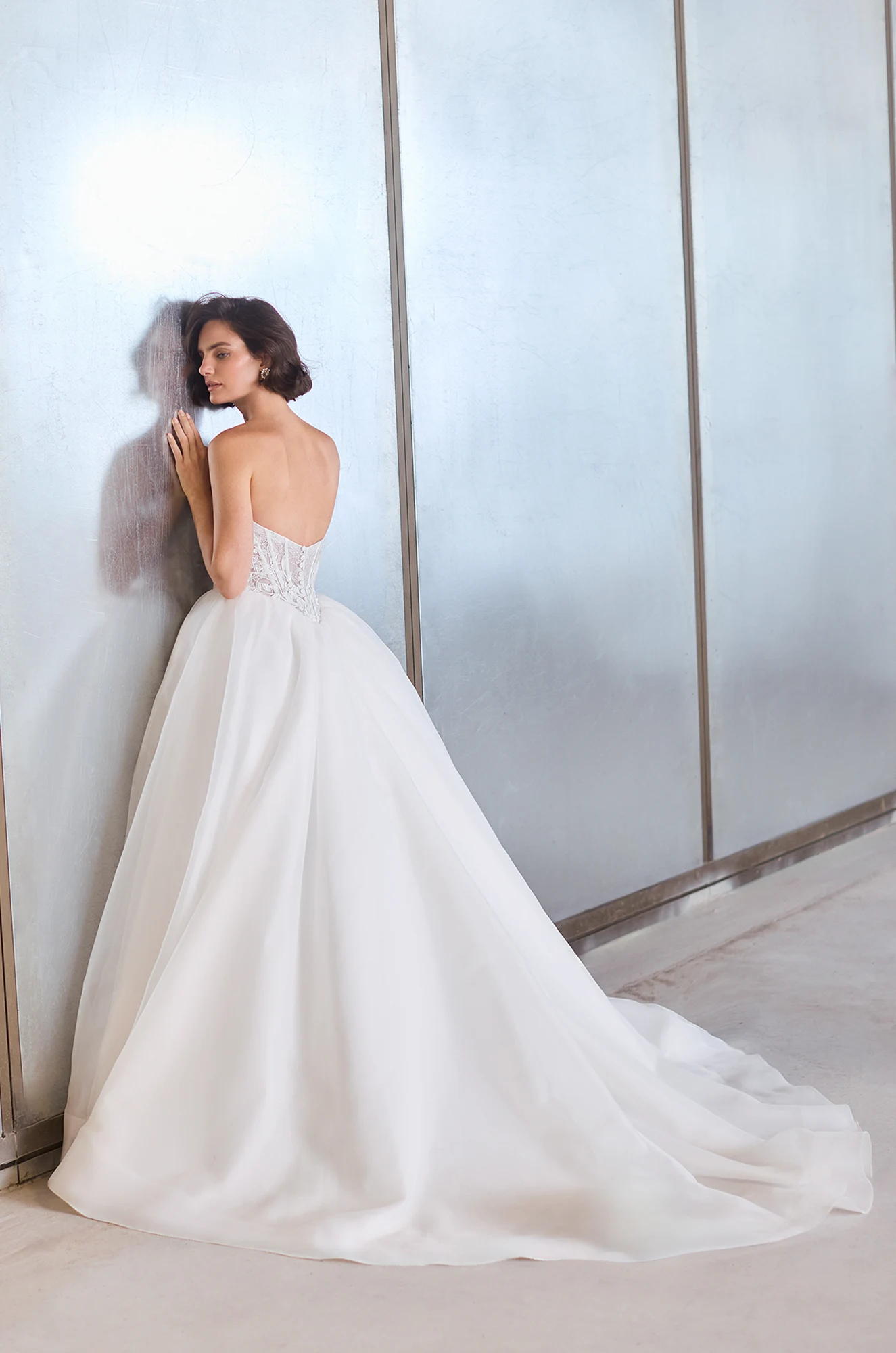 Back view of a strapless sheer Lace bodice wedding dress with a basque waistline and a gathered Organza ball gown skirt, in front of a wall.
