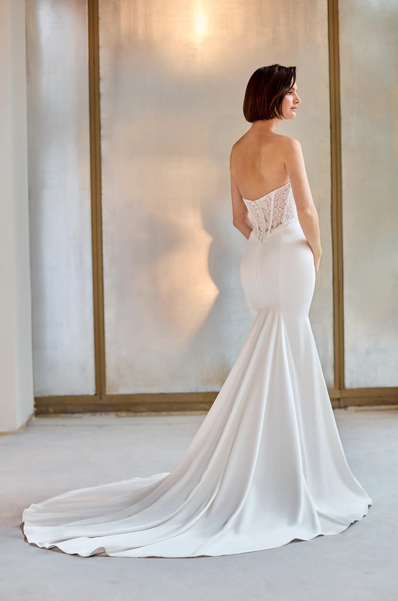 Back view of a strapless wedding dress with a sheer Lace bodice with buttons down the back bodice, and a fit and flare Charmeuse skirt, in front of a wall.