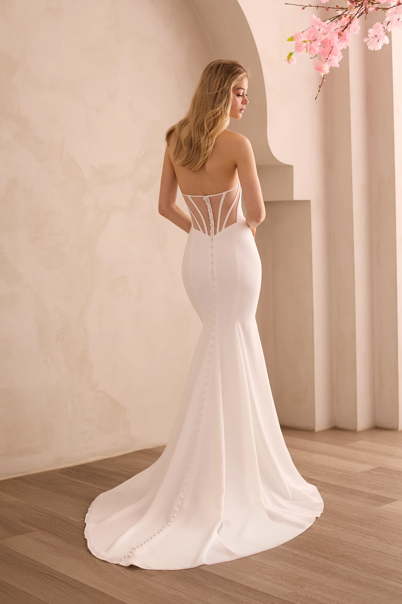 Back view of a Crepe wedding dress with a sheer tulle back with curved boning and buttons, with a fit and flare skirt, in front of a decorative wall.
