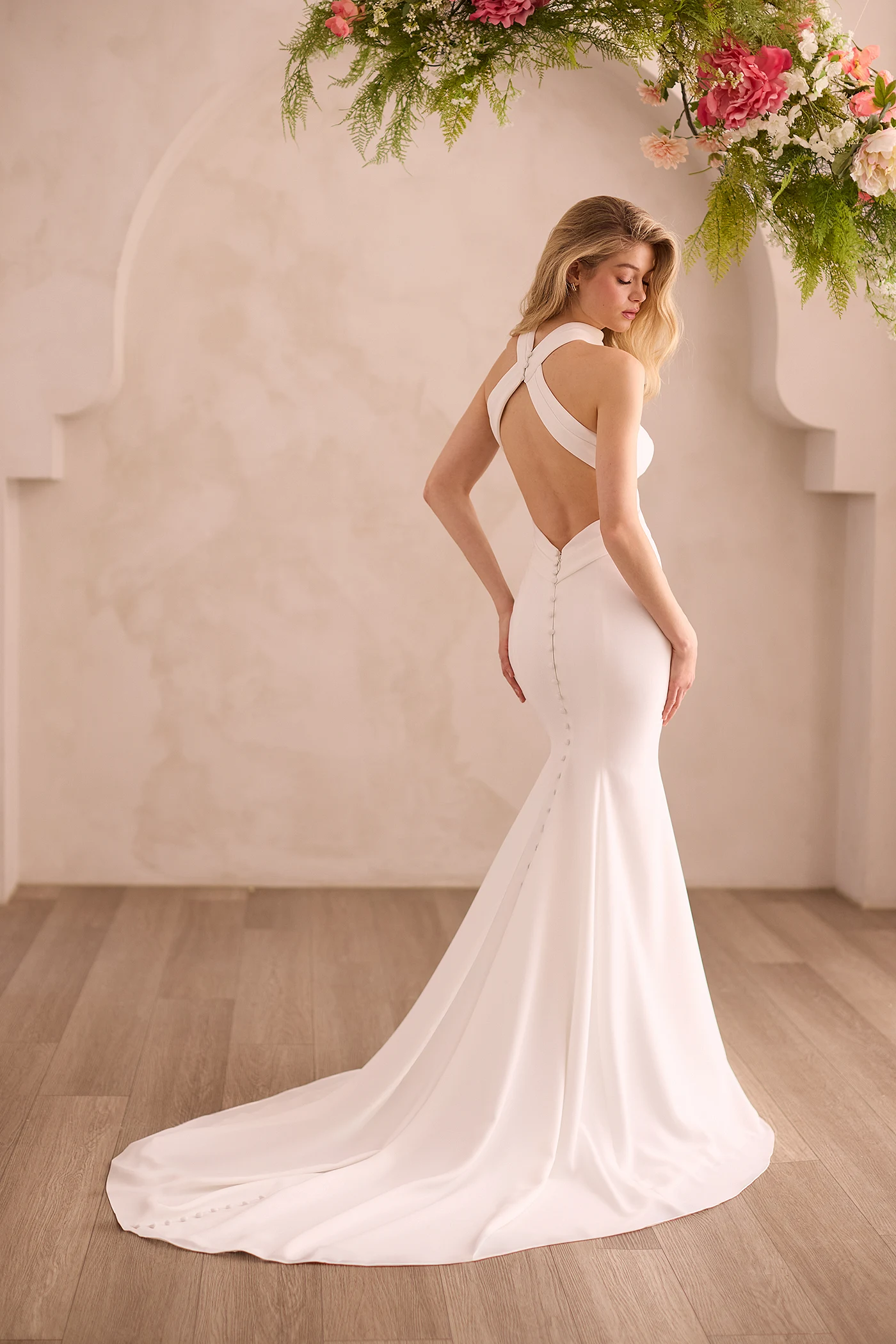 Back view of a sleeveless Crepe wedding dress with pleated straps crossing over the back and buttons down the train of a fit and flare skirt, in front of a decorative wall.
