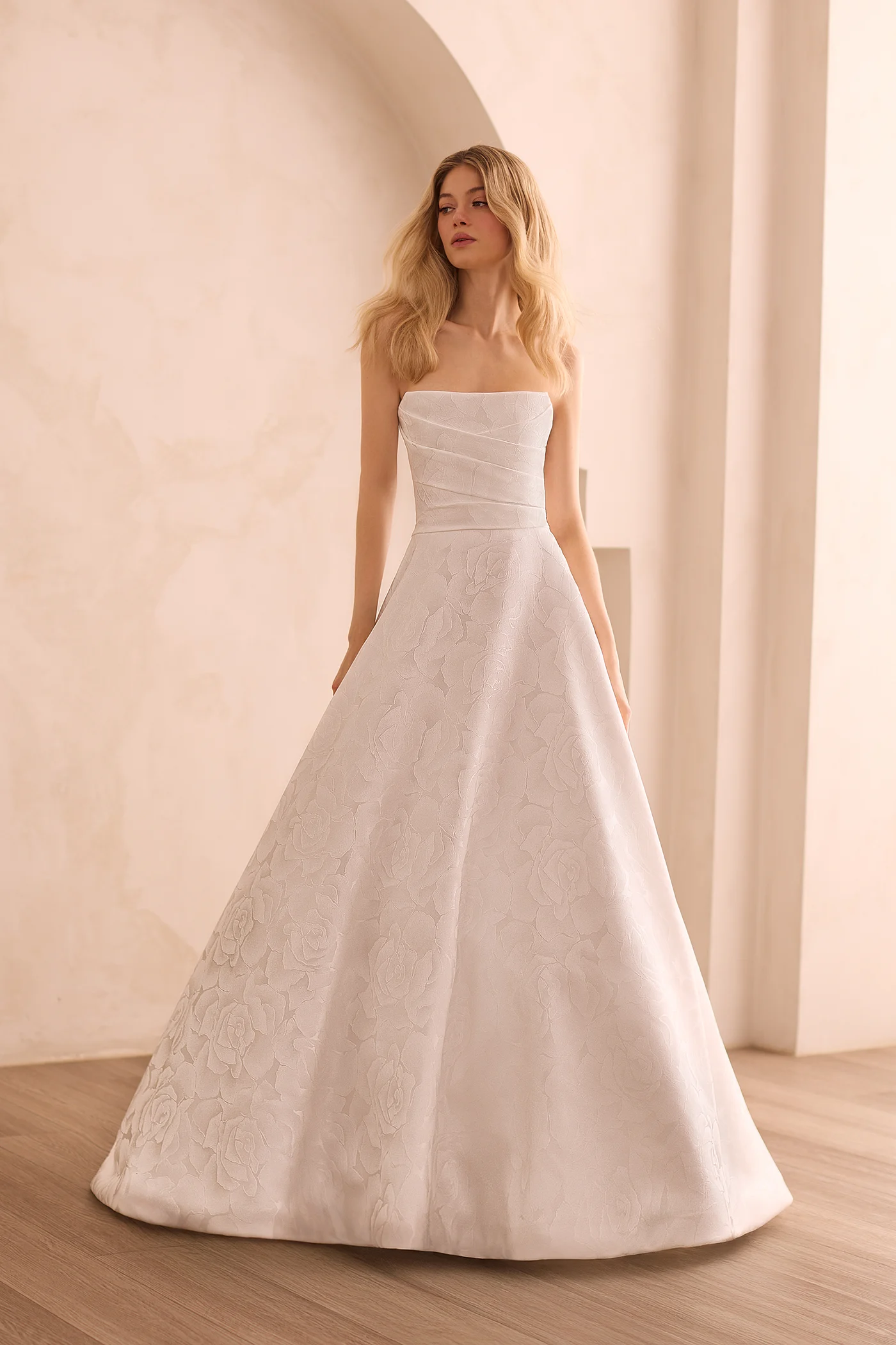 Front view of a strapless Jacquard wedding dress with pleated bodice, scoop neckline, and an A-line skirt in front of a decorative wall.
