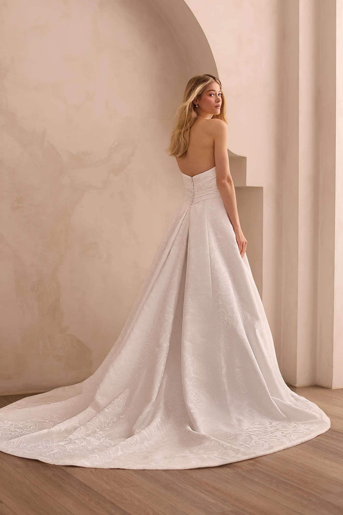 Back view of a strapless Jacquard wedding dress with buttons on the back of the bodice and pleats on the back of an A-line skirt, in front of a decorative wall.