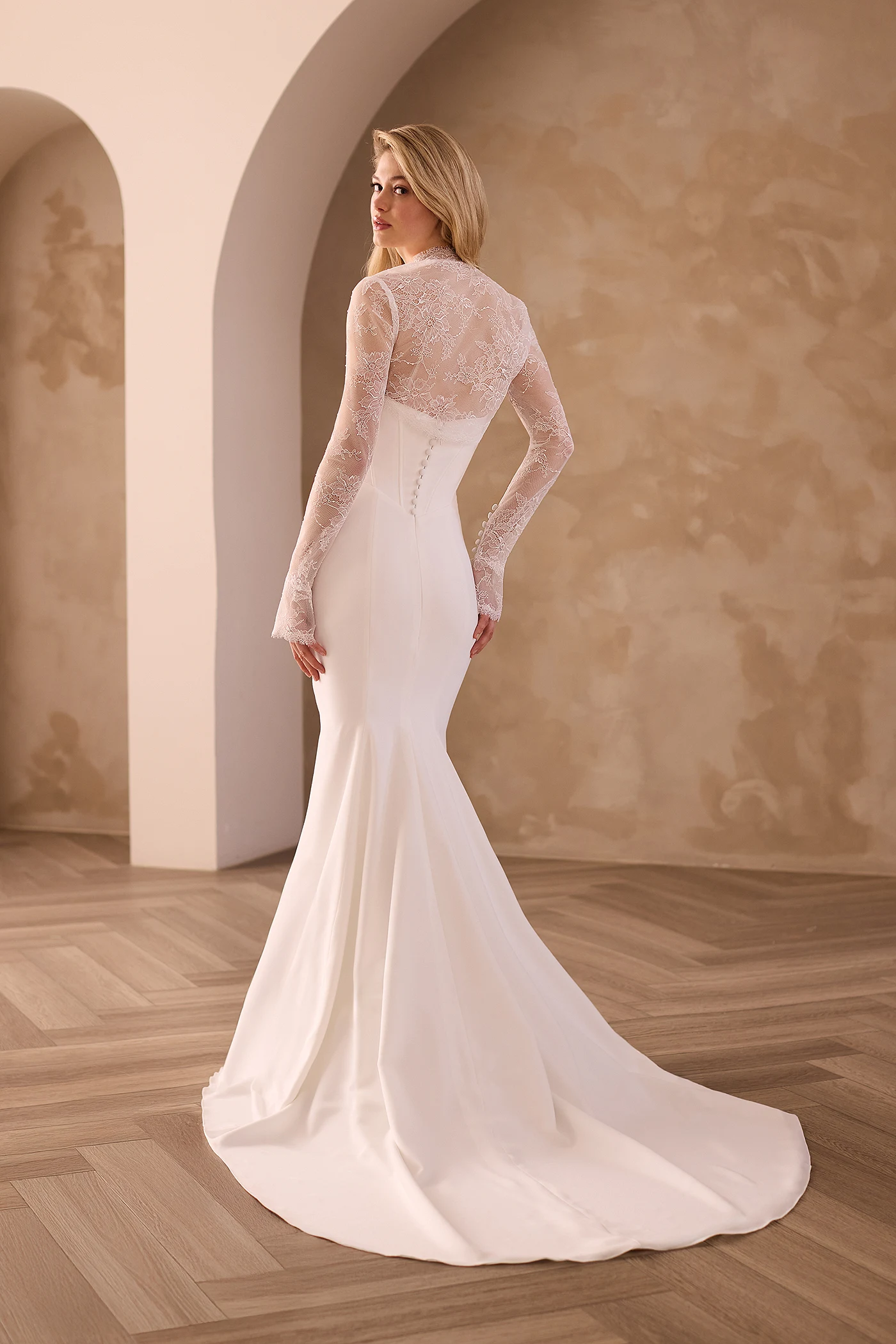 Back view of a Crepe wedding dress with buttons down the back of the bodice and a fit and flare Crepe skirt and a long sleeve Lace bolero, in front of an archway.