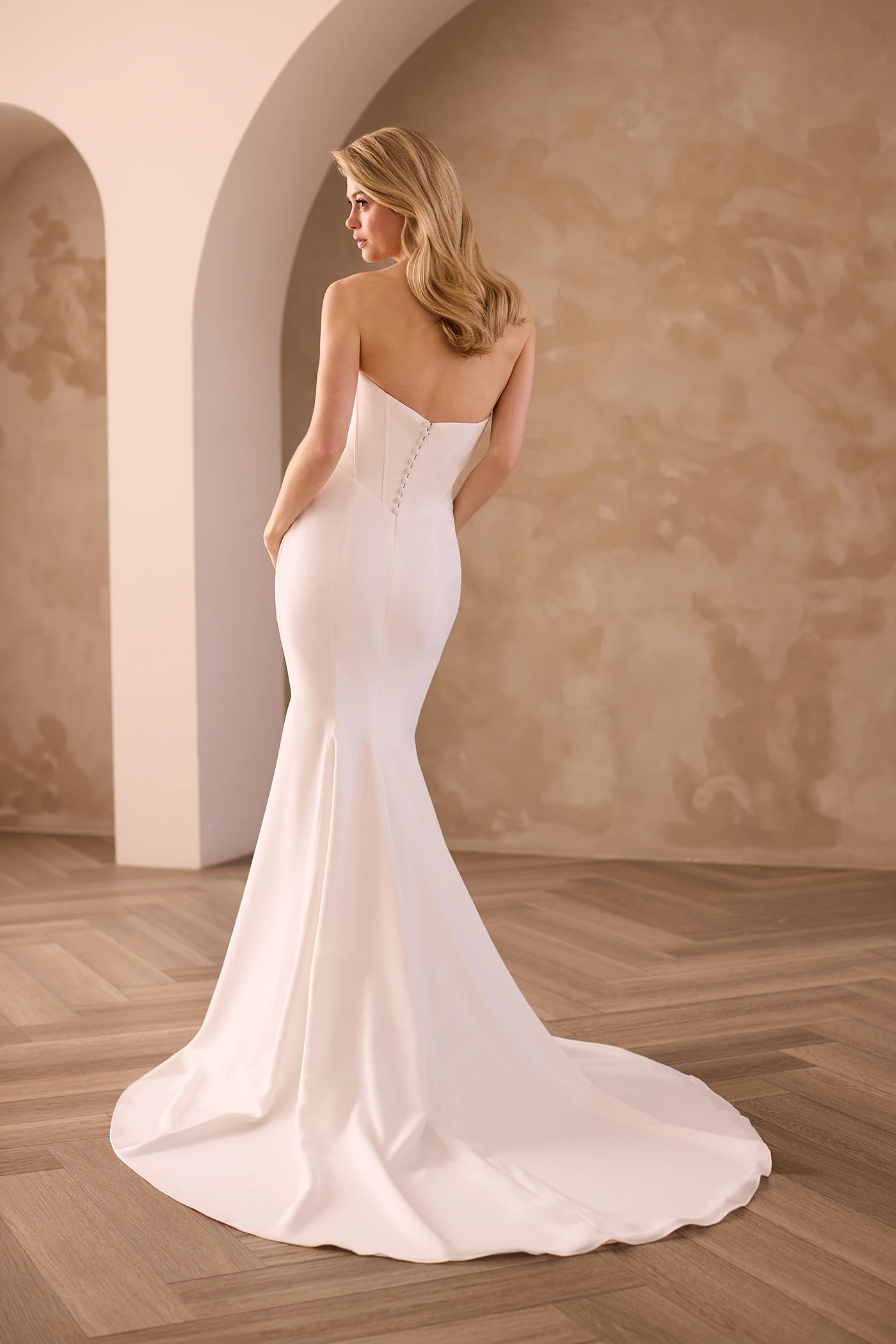 Back view of a strapless Crepe wedding dress with buttons down the back of the bodice and a fit and flare Crepe skirt, in front of an archway.