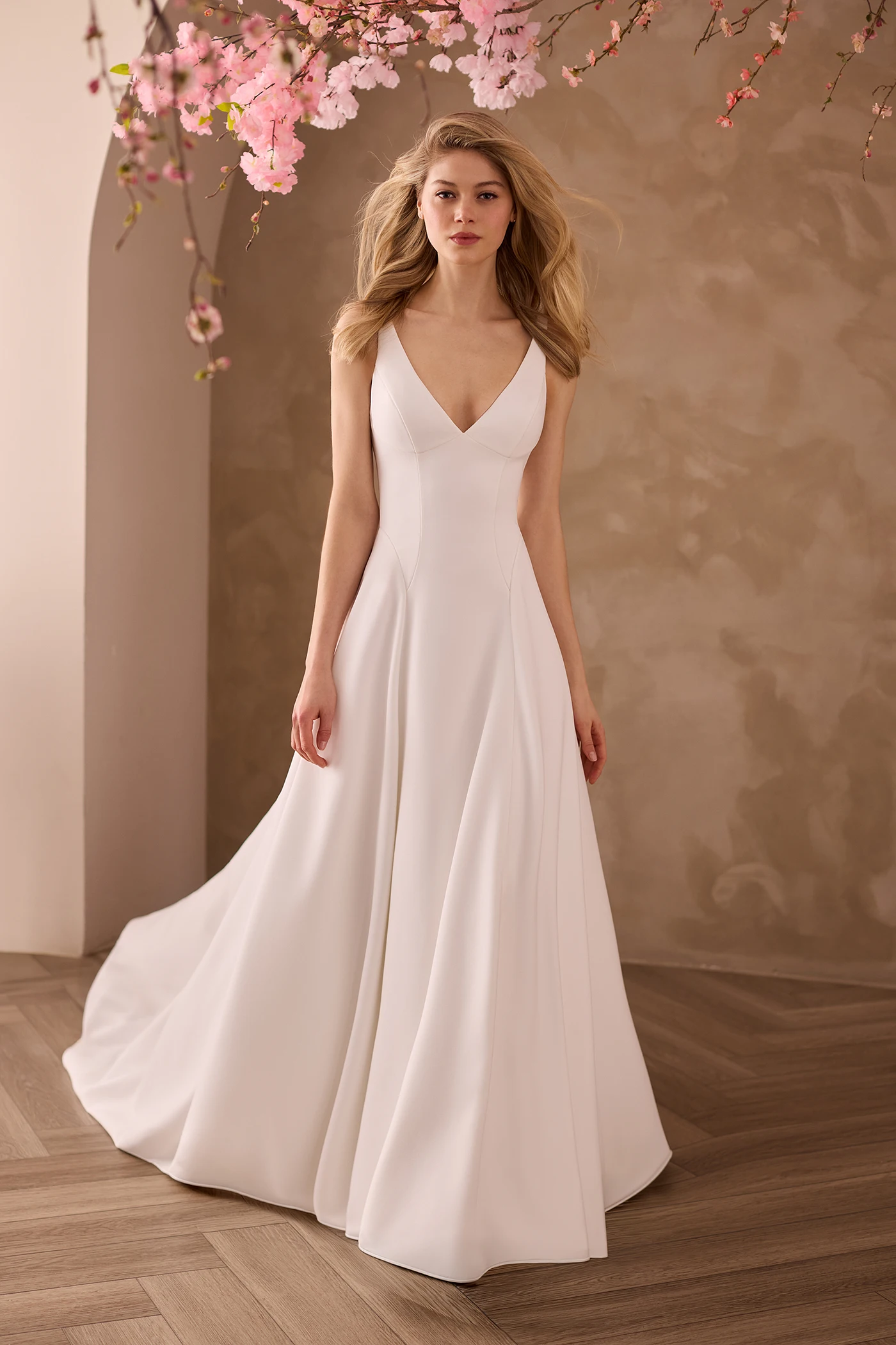 Front view of a sleeveless Crepe V-neckline wedding dress with princess seams and a full A-line skirt, in front of an archway with flowers above the model.
