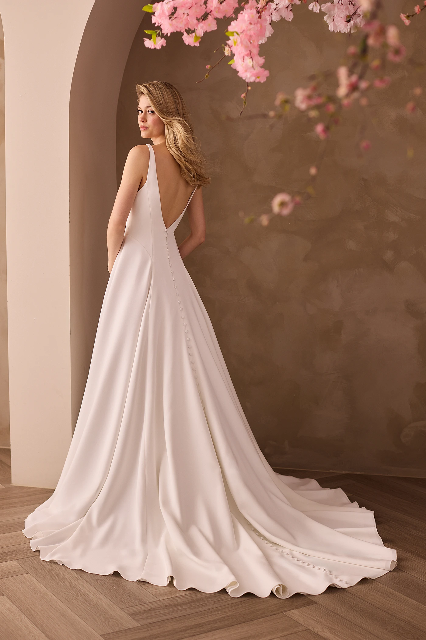 Back view of a sleeveless Crepe wedding dress with buttons going down the back of the dress to the end of the train on an A-line skirt, in front of an archway.