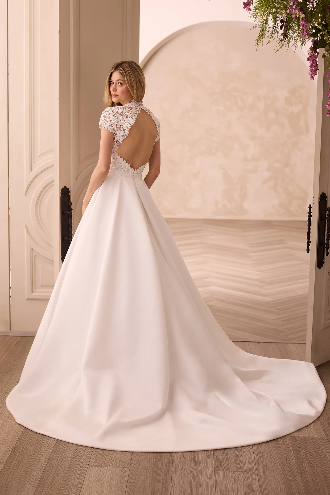 Back view of a wedding dress with a Lace keyhole back and cap sleeves on the bodice and a Satin A-line skirt in front of a double door entrance.