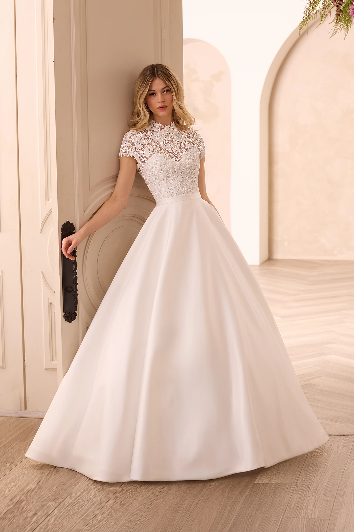 Front view of a wedding dress with a Lace cap sleeve bodice and an A-line Satin skirt, in front of a double door entrance.