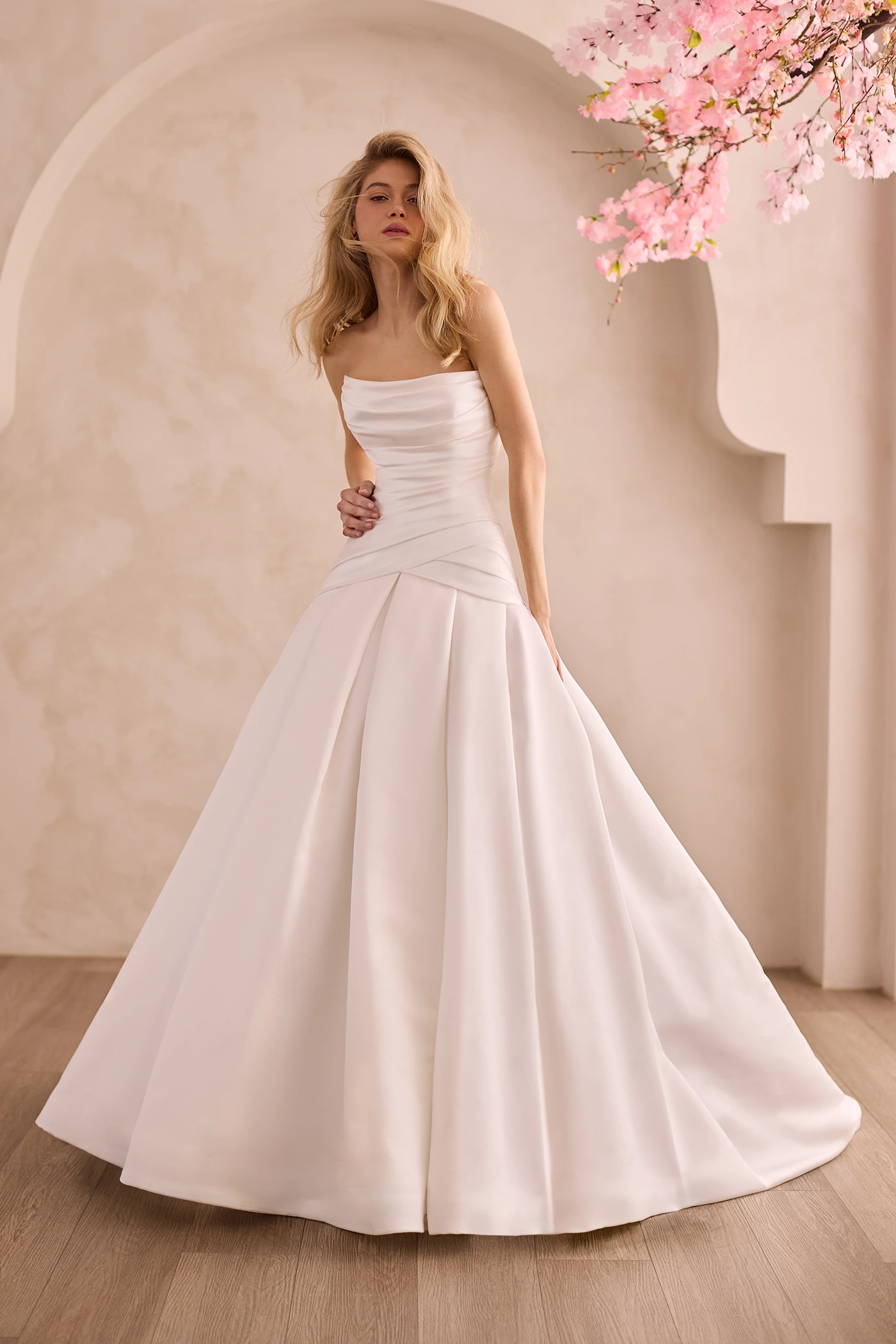 Front view of a strapless Satin wedding dress with pleated drop waisted bodice and scoop neckline, with a box pleated skirt in front of a decorative wall and flowers.