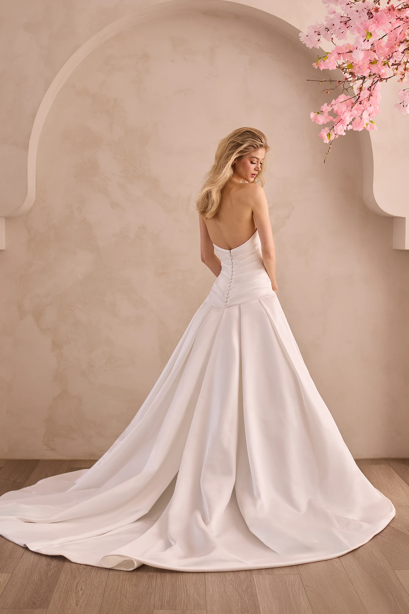 Back view of a strapless Satin wedding dress with pleated drop waisted bodice and buttons down back of bodice, with a box pleated skirt in front of a decorative wall and flowers.