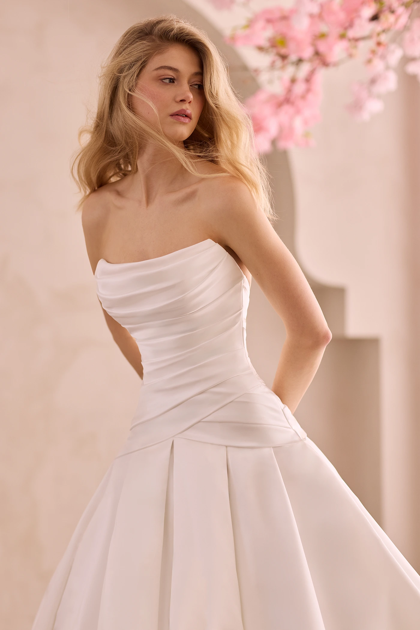 Close up view of a strapless Satin wedding dress with pleated drop waisted bodice and scoop neckline, with a box pleated skirt in front of a decorative wall and flowers.