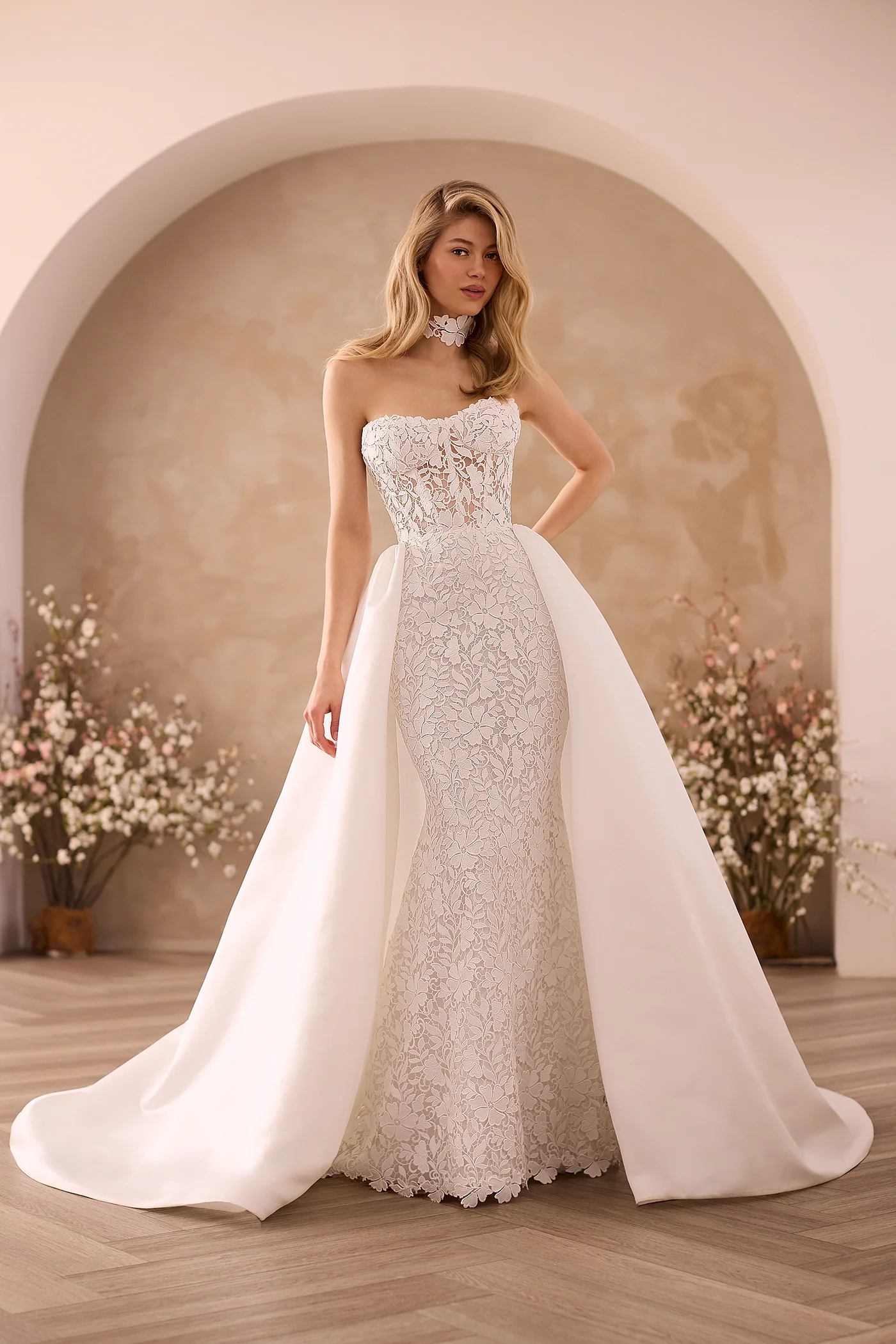 Front view of a strapless Lace wedding dress with a sheer Lace bodice and fit and flare skirt, a detachable Satin train and a Lace choker, in front of an archway and flower planters.