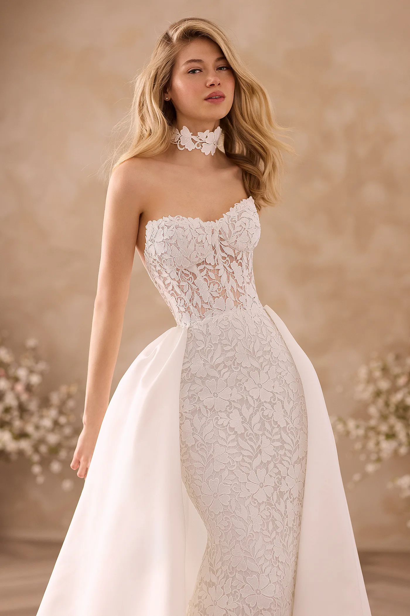Close up view of a strapless Lace wedding dress with a sheer Lace bodice and a fitted skirt, a detachable Satin train and a Lace choker, in front of a wall and flowers.