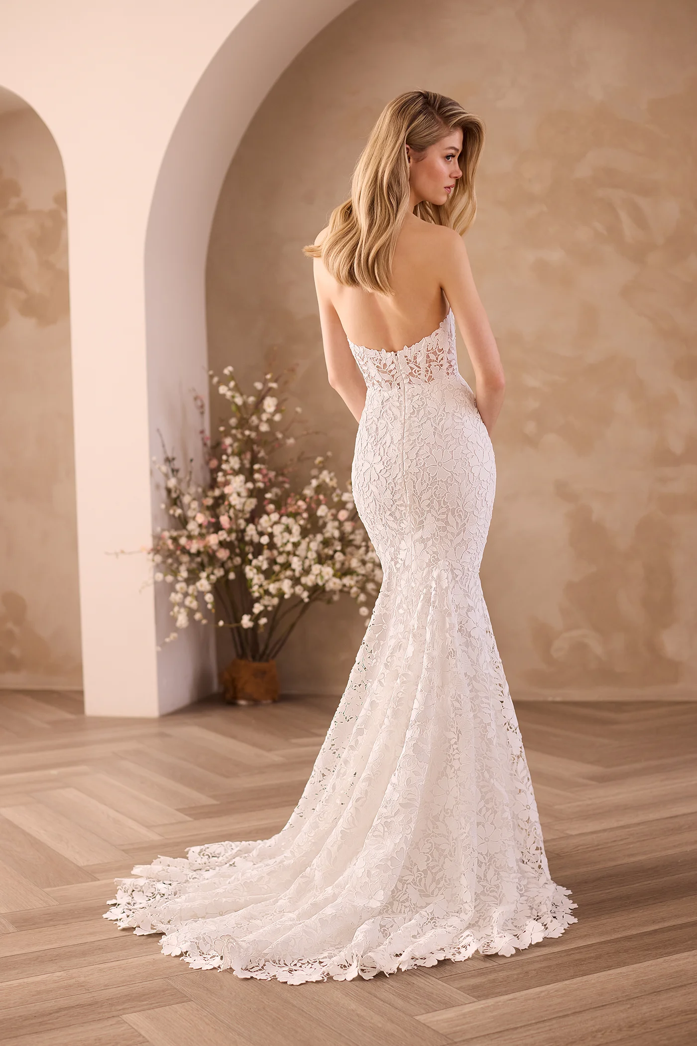 Back view of a strapless Lace wedding dress with a sheer Lace bodice and a fit and flare skirt, in front of an archway and flower planters.