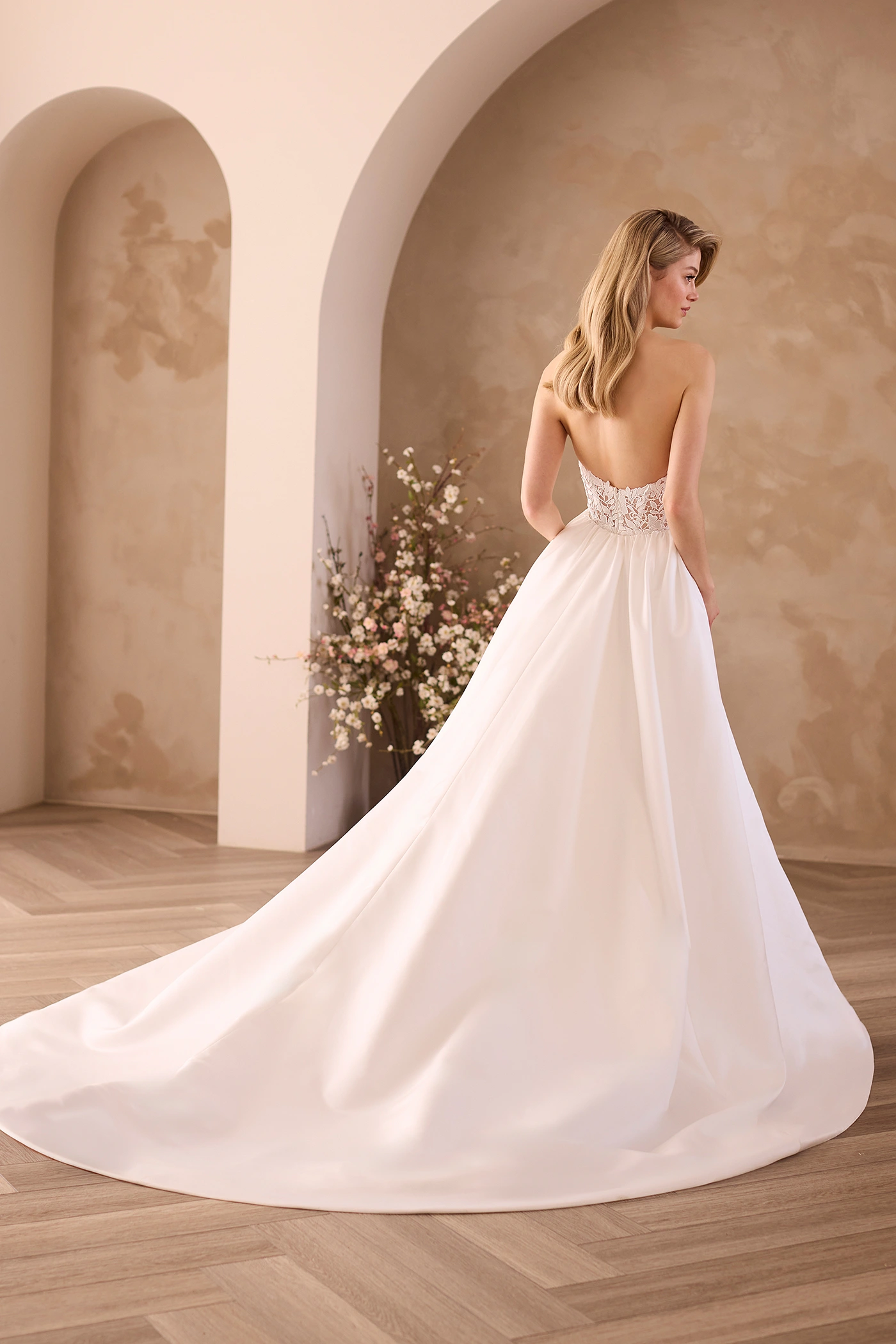 Back view of a strapless wedding dress with a sheer Lace bodice and a detachable Satin train, in front of archways and a flower planter.