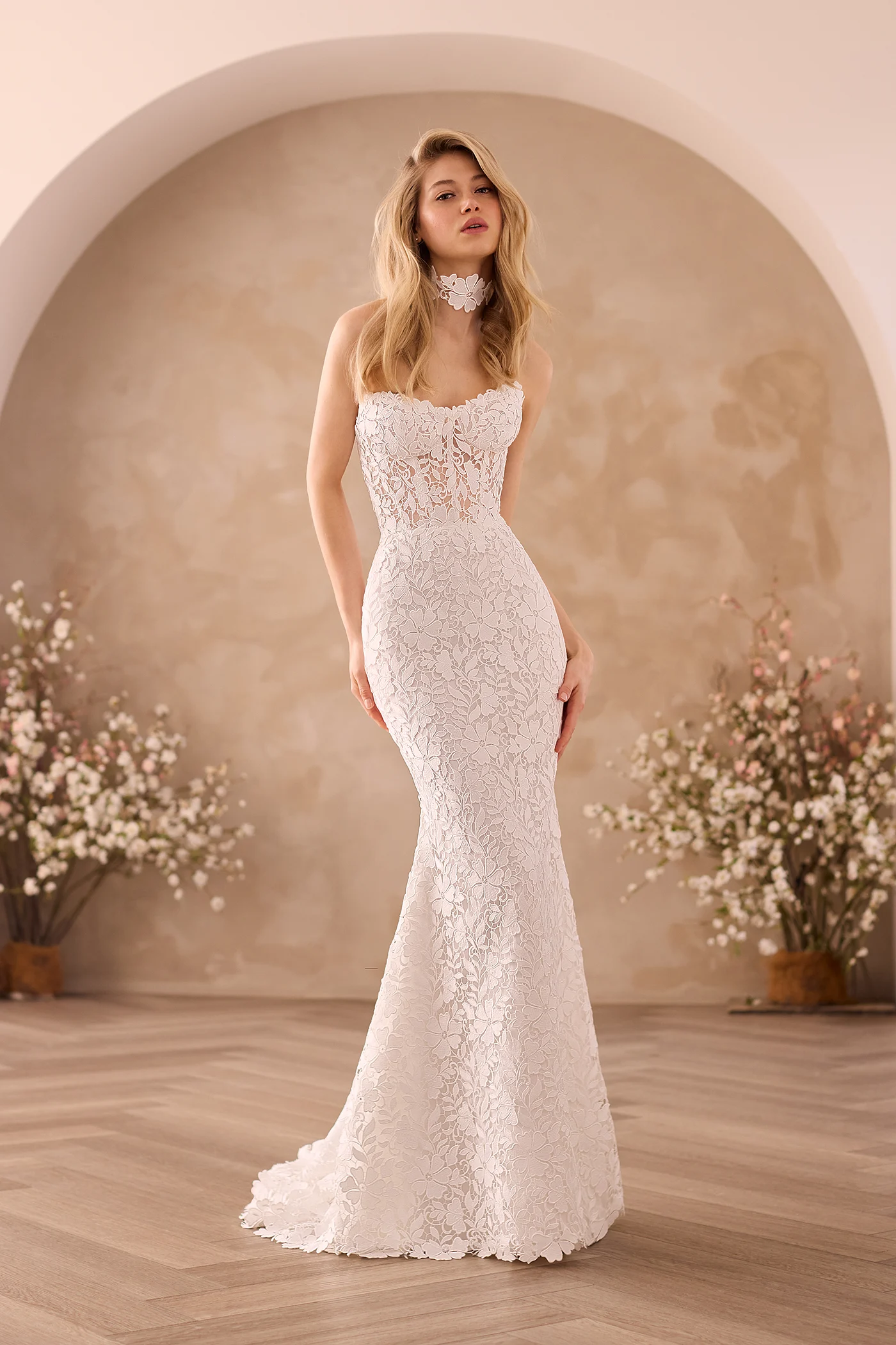 Front view of a strapless Lace wedding dress with a sheer Lace bodice and a fit and flare skirt, with a Lace choker in front of an archway with flower planters.