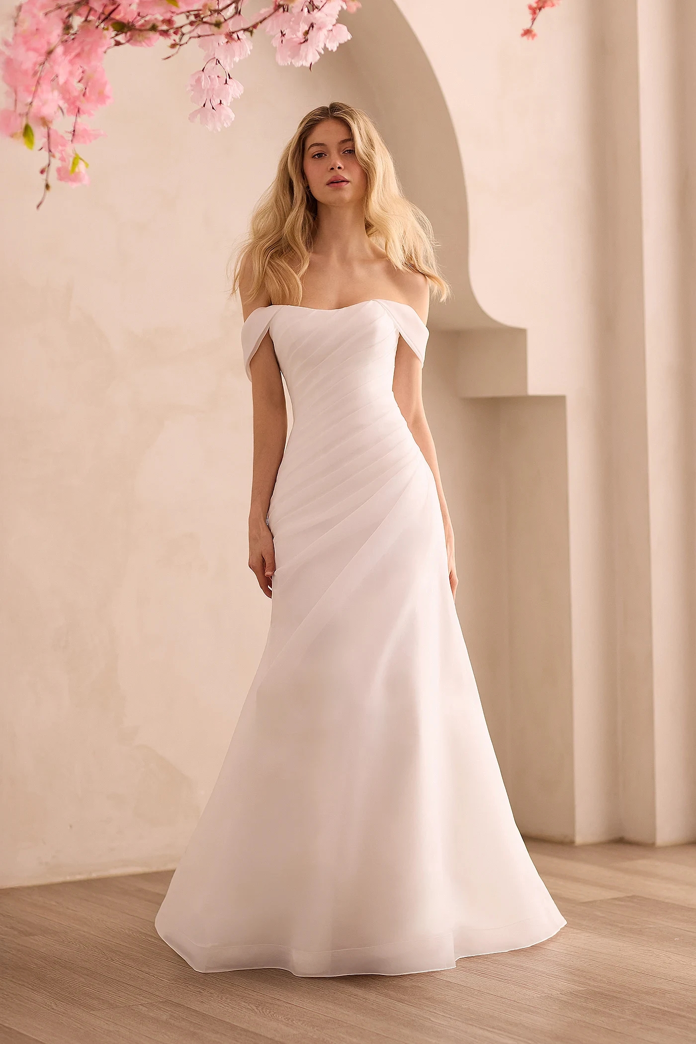 Front view of an Organdy wedding dress with asymmetric pleated bodice, draped sleeves, and a draped A-line skirt, in front of a decorative wall.