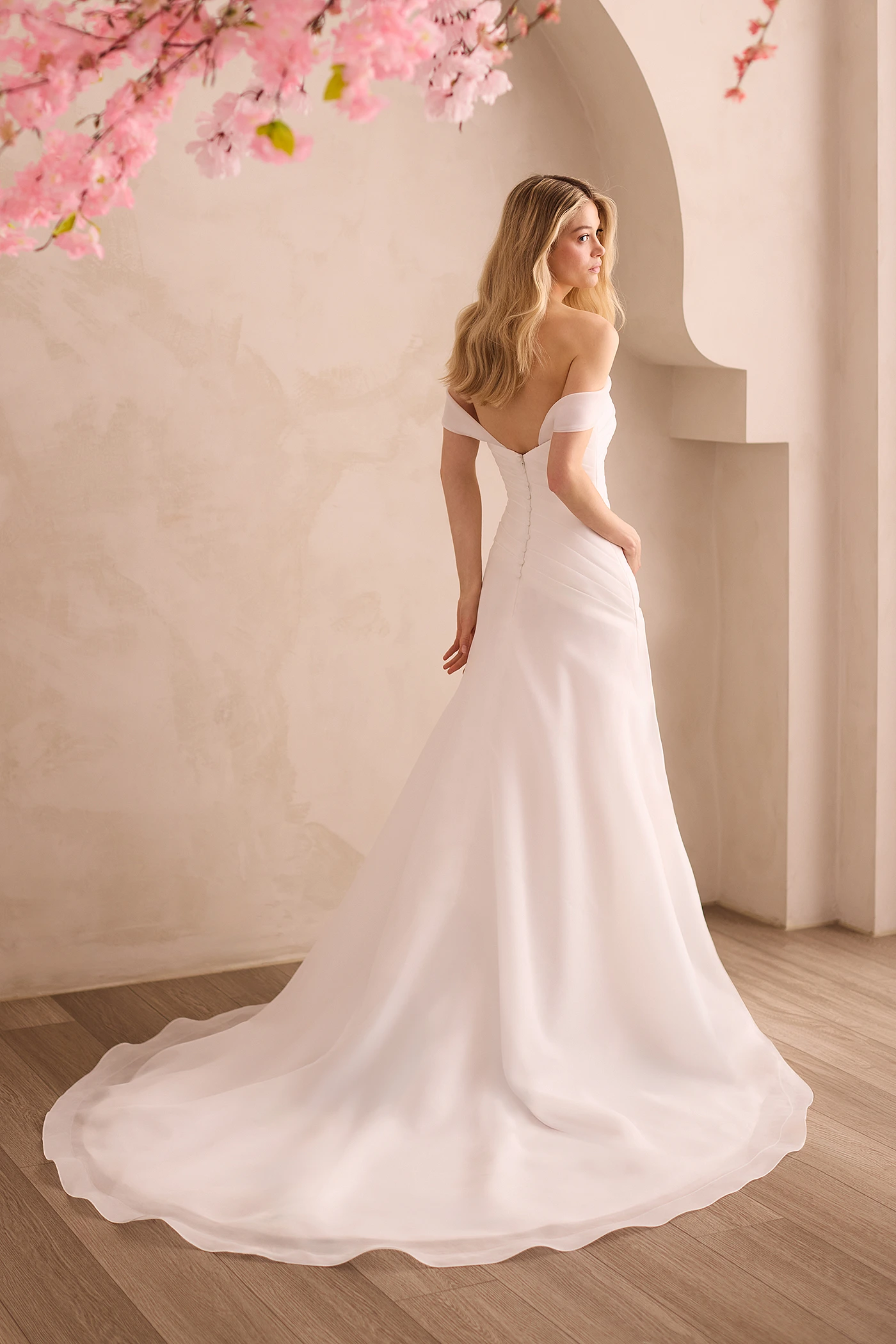 Back view of an Organdy wedding dress with pleated bodice and A-line skirt, in front of a decorative wall with flowers above the model.