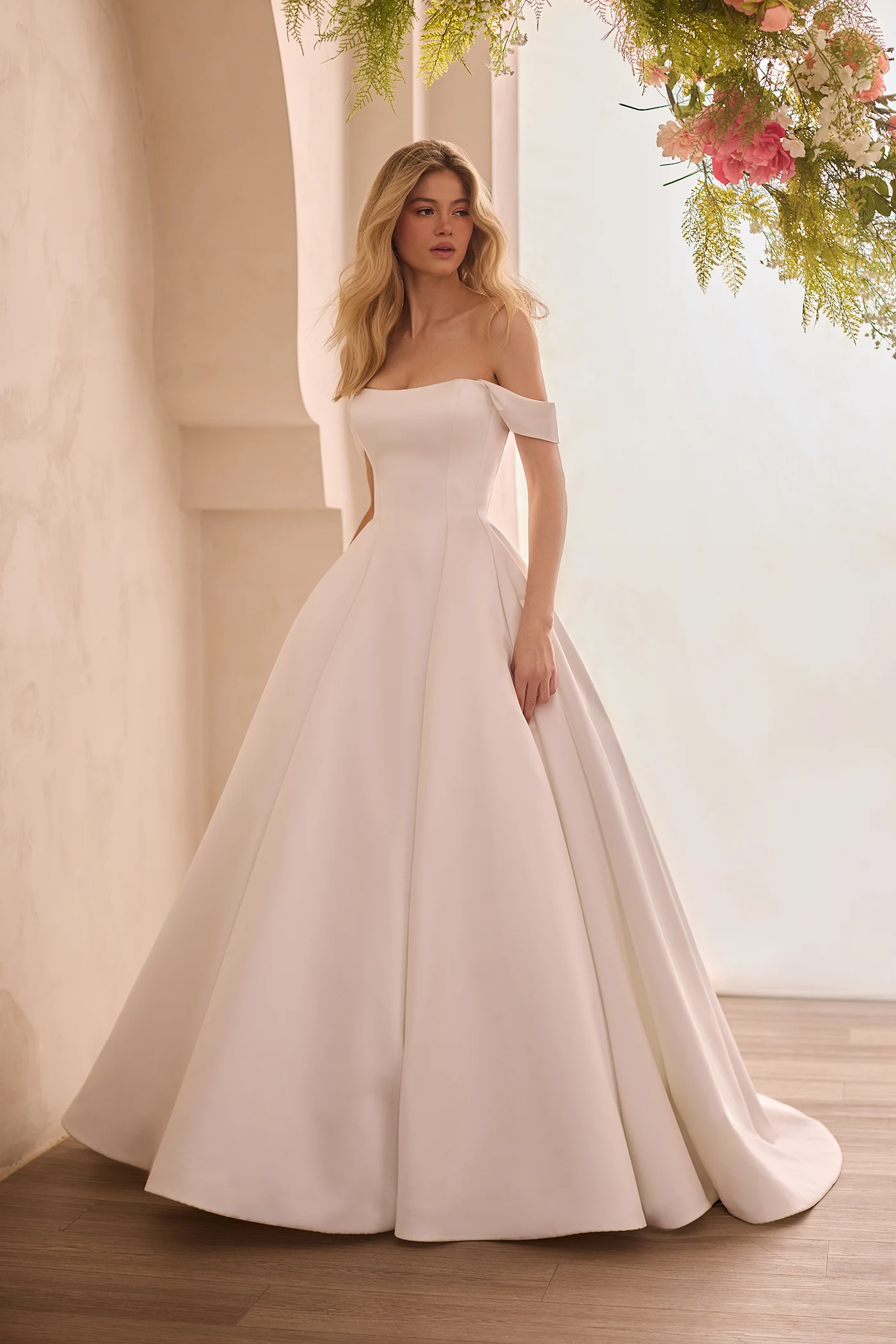 Front view of a Satin princess seam wedding dress with scoop neckline and detachable draped sleeves with a full A-line skirt, in front of a decorative wall with flowers above the model.