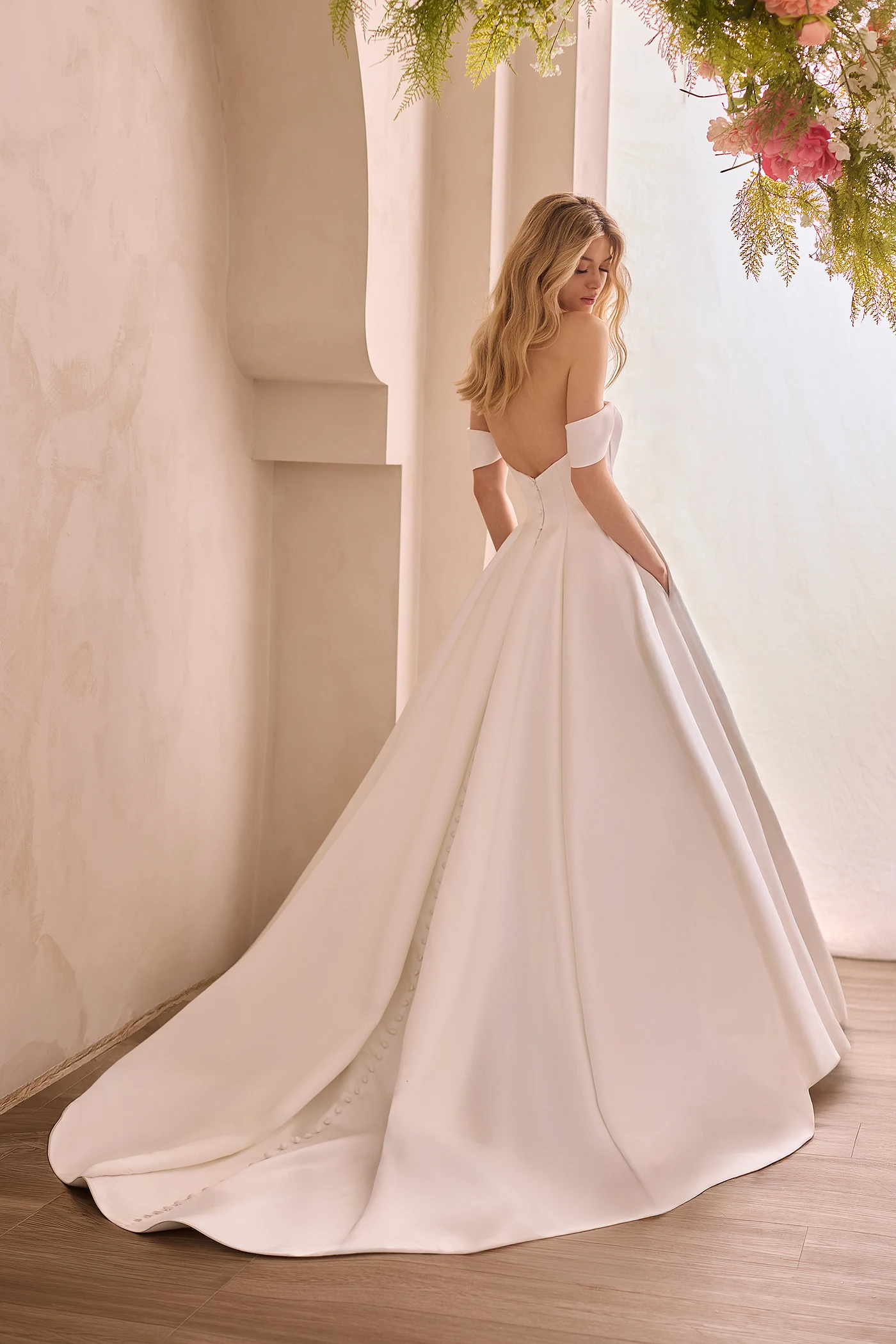 Back view of a Satin wedding dress with draped sleeves and a full A-line skirt with pockets and buttons down the back of the dress to the hem, in front of a decorative wall with flowers above the model.