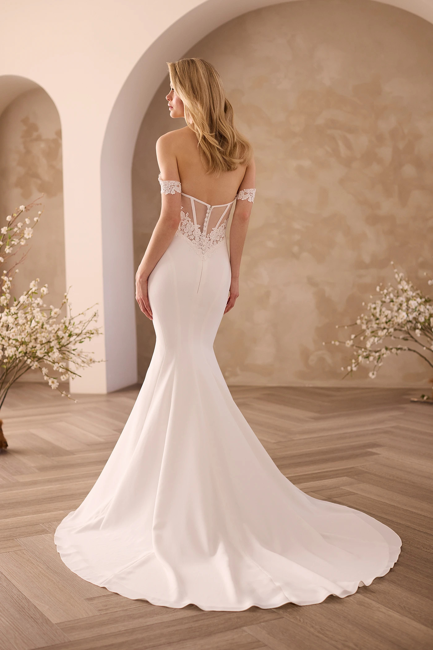 Back view of a wedding dress with Lace draped sleeves and a sheer tulle back with Lace appliques along lower back, with a fit and flare Crepe skirt, in front of archways.