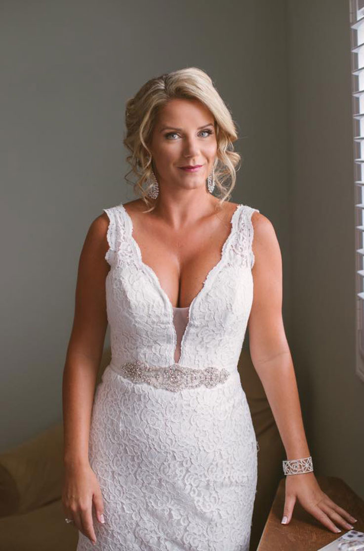 Close-up of bride in a scalloped plunging neck wedding dress with lace and a beaded waistband, posing indoors by a window.
