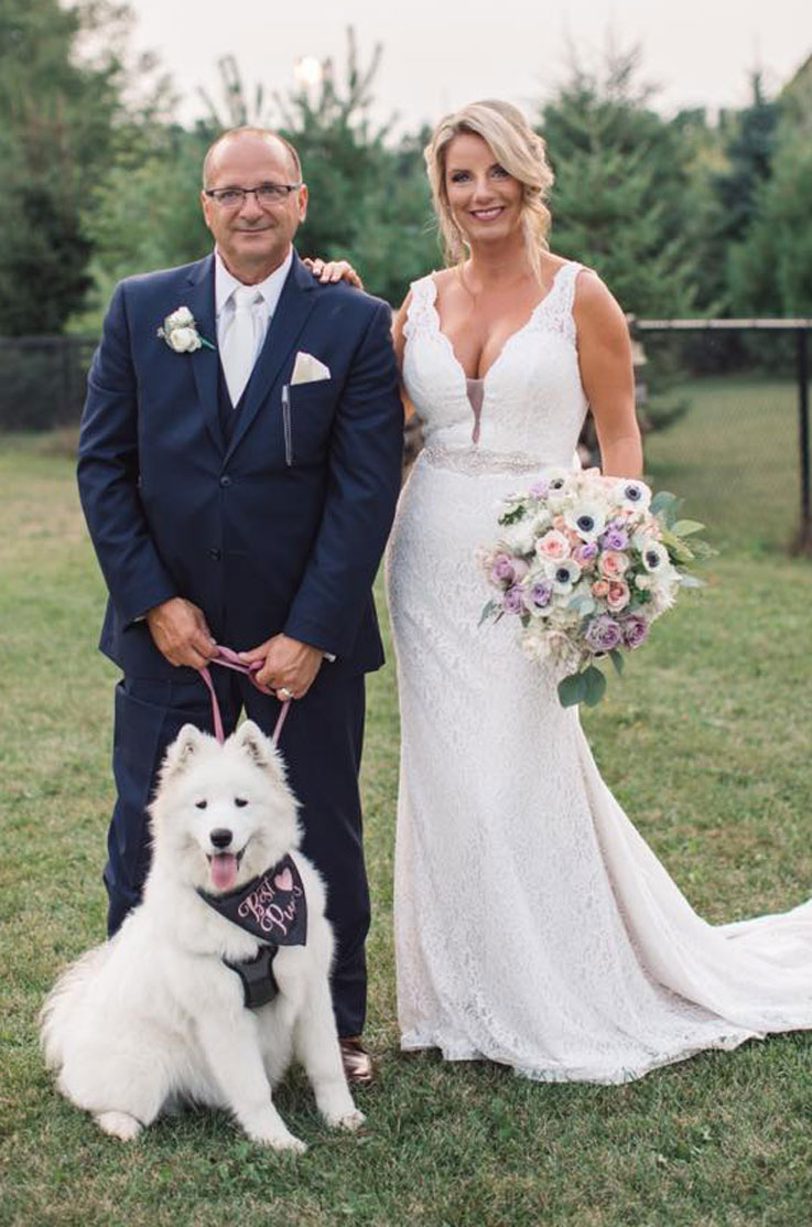 Bride in a plunging neck lace gown with a beaded belt, smiles and poses next to groom in a navy suit outdoors with their dog.