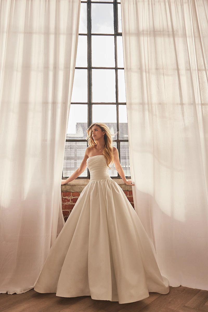 Strapless ballgown wedding dress with a ruched bodice and full satin skirt, shown in front of tall industrial windows.