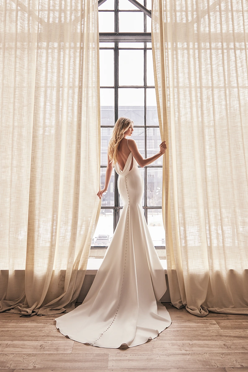 Sleeveless crepe wedding dress with a low draped back, covered buttons, and long flowing train in front of a tall window.