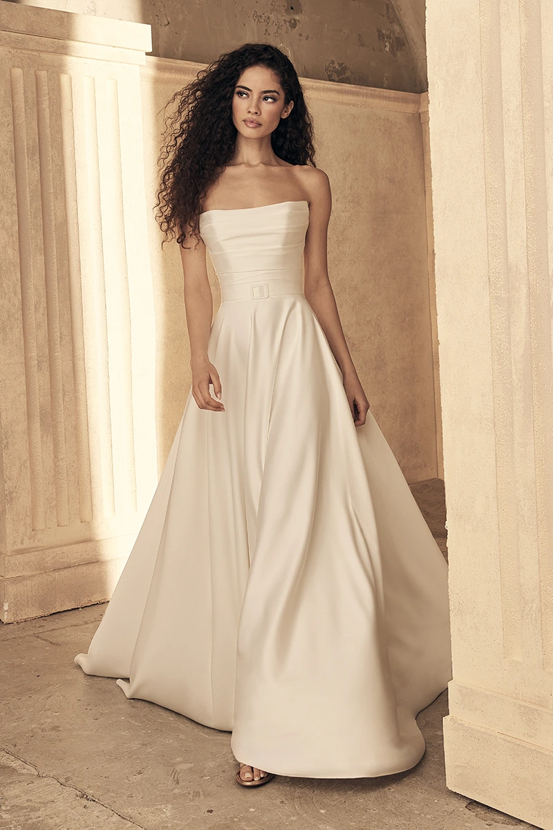 Strapless A-line gown with a straight neckline, structured pleated bodice, slim satin belt, and flowing full skirt.